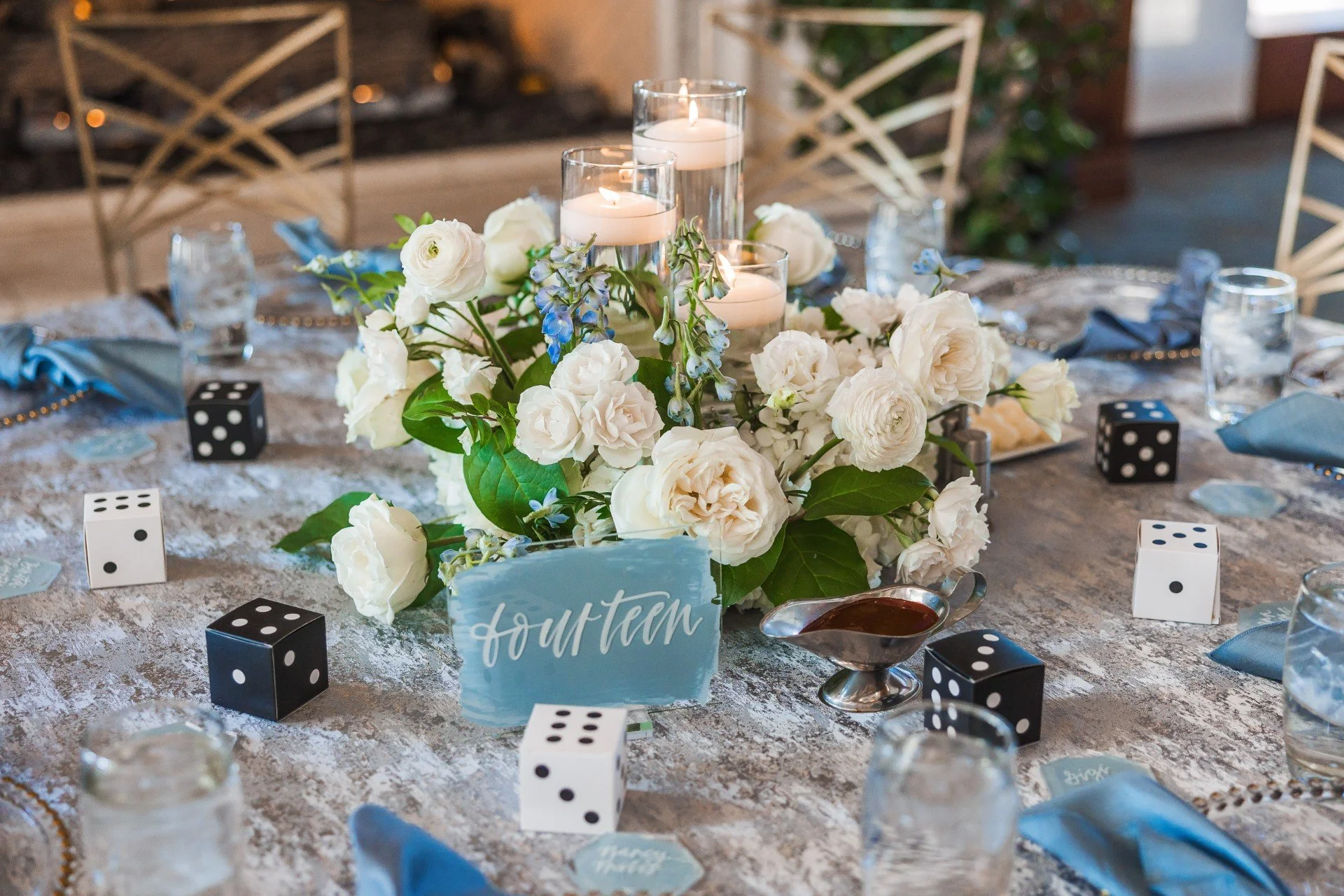 A decorated table with a floral centerpiece of white roses, blue flowers, and green leaves, surrounded by black and white dice, blue folded napkins, candles in glass holders, and a small sign saying 'Table Four' for a wedding.