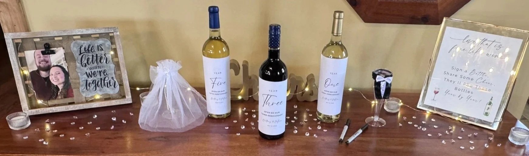 Table decorated with wine bottles, a framed photo of a couple, signs, markers, and decorative fairy lights for a celebration or wedding, with scattered small decorations on the table.