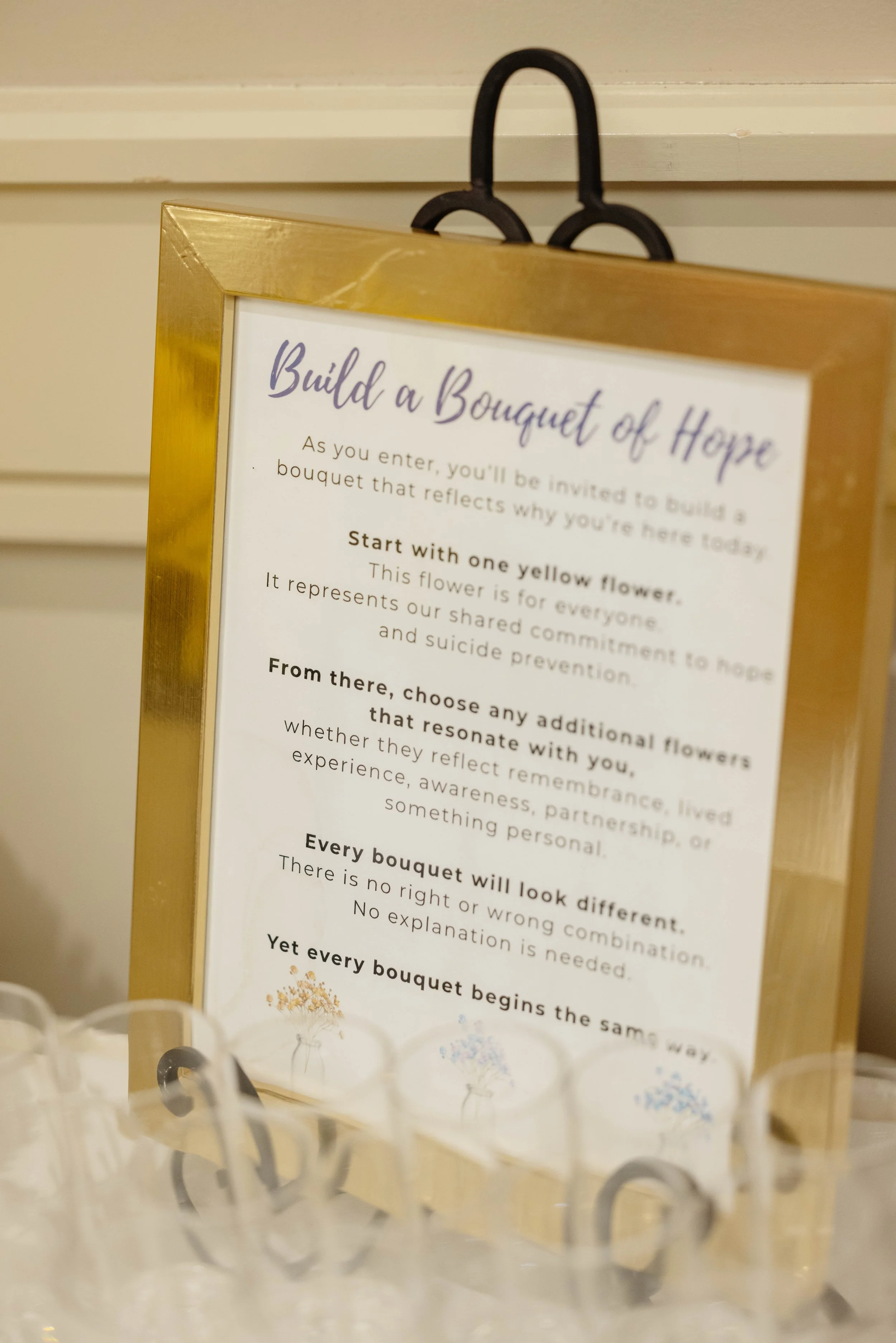 A framed sign with gold border on display with a partially visible floral arrangement in the foreground. The sign contains text about building a bouquet of hope.