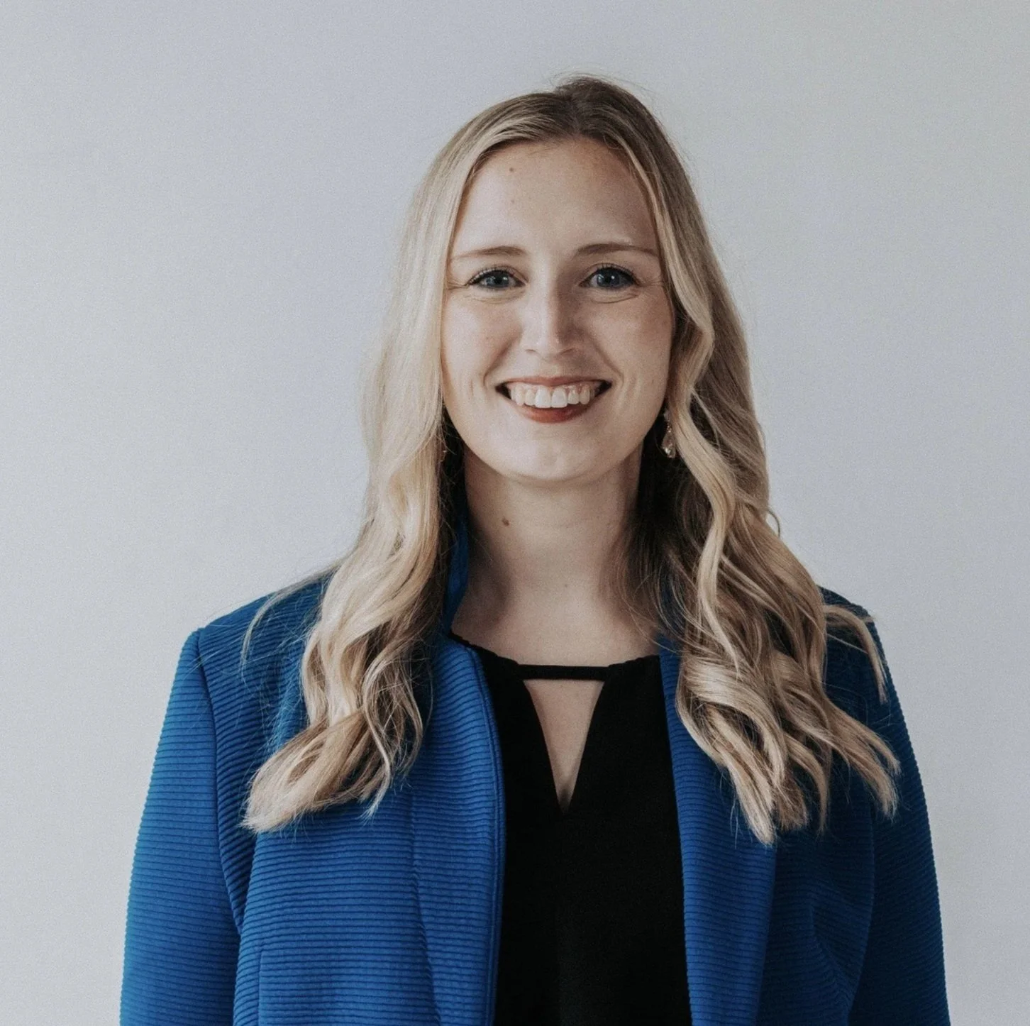 A woman with blonde wavy hair wearing a blue blazer and black top, smiling against a plain white wall.