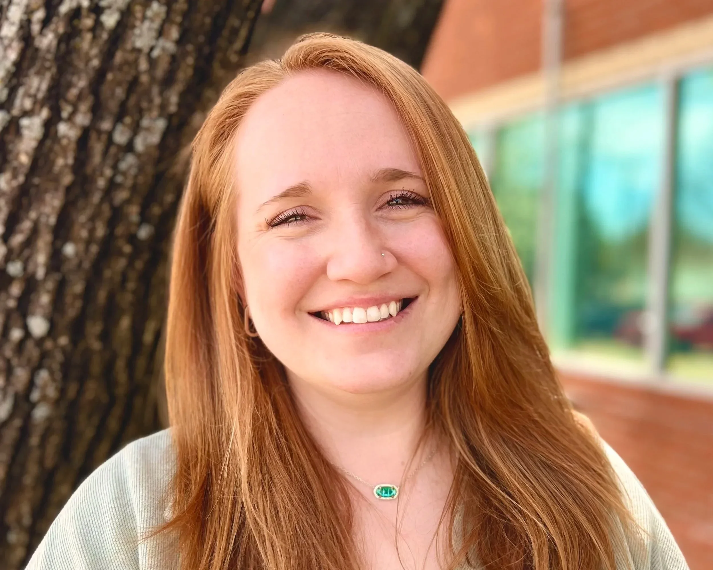 A smiling woman with red hair, wearing a necklace, standing outside near a tree with a building with large windows in the background.