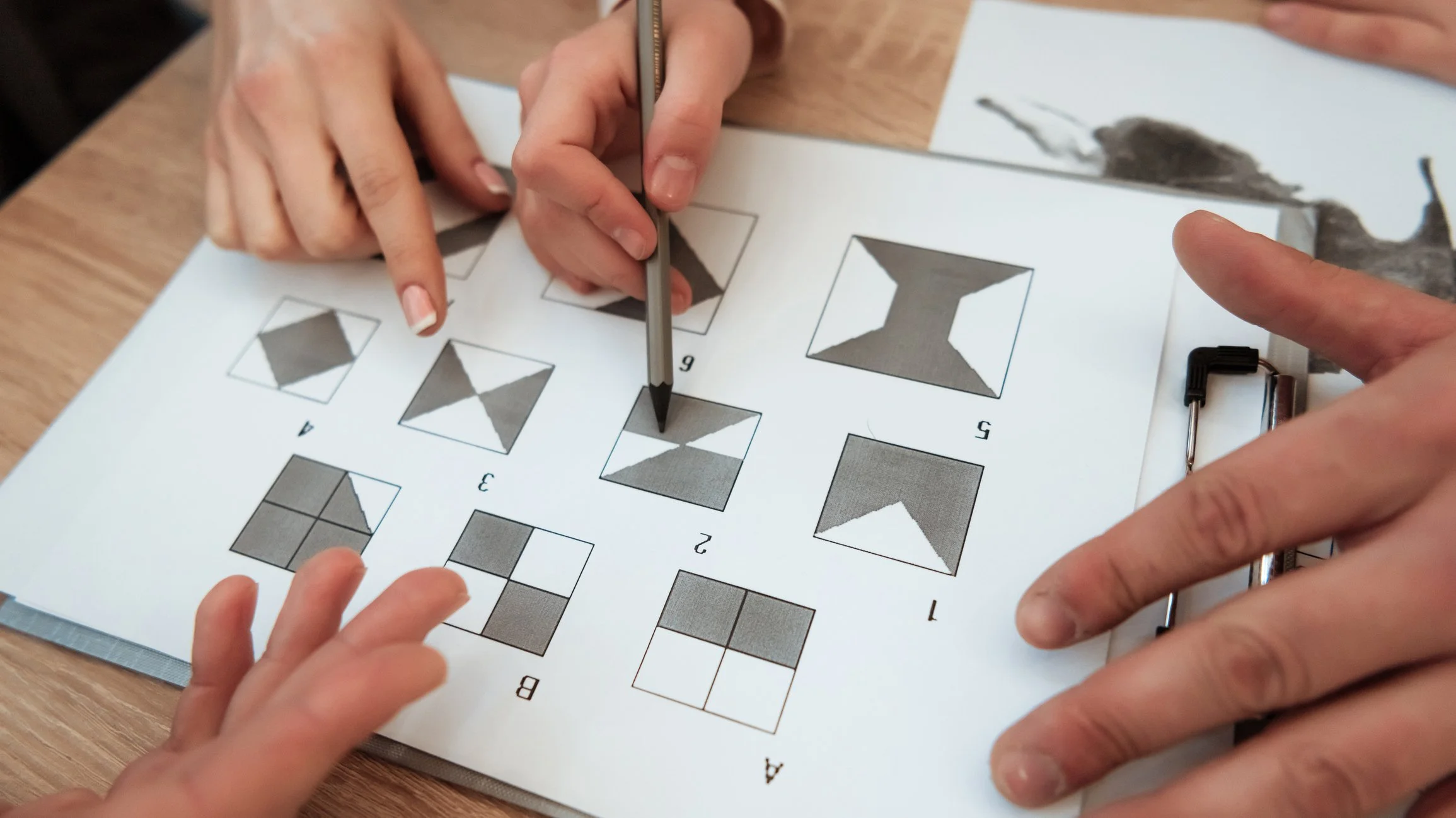 People working on a workbook with geometric pattern puzzles, using a pen and a sharpener on a wooden table.