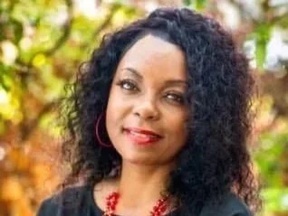 Portrait of a woman with curly black hair, wearing red earrings and a necklace, outdoors with autumn leaves in the background.