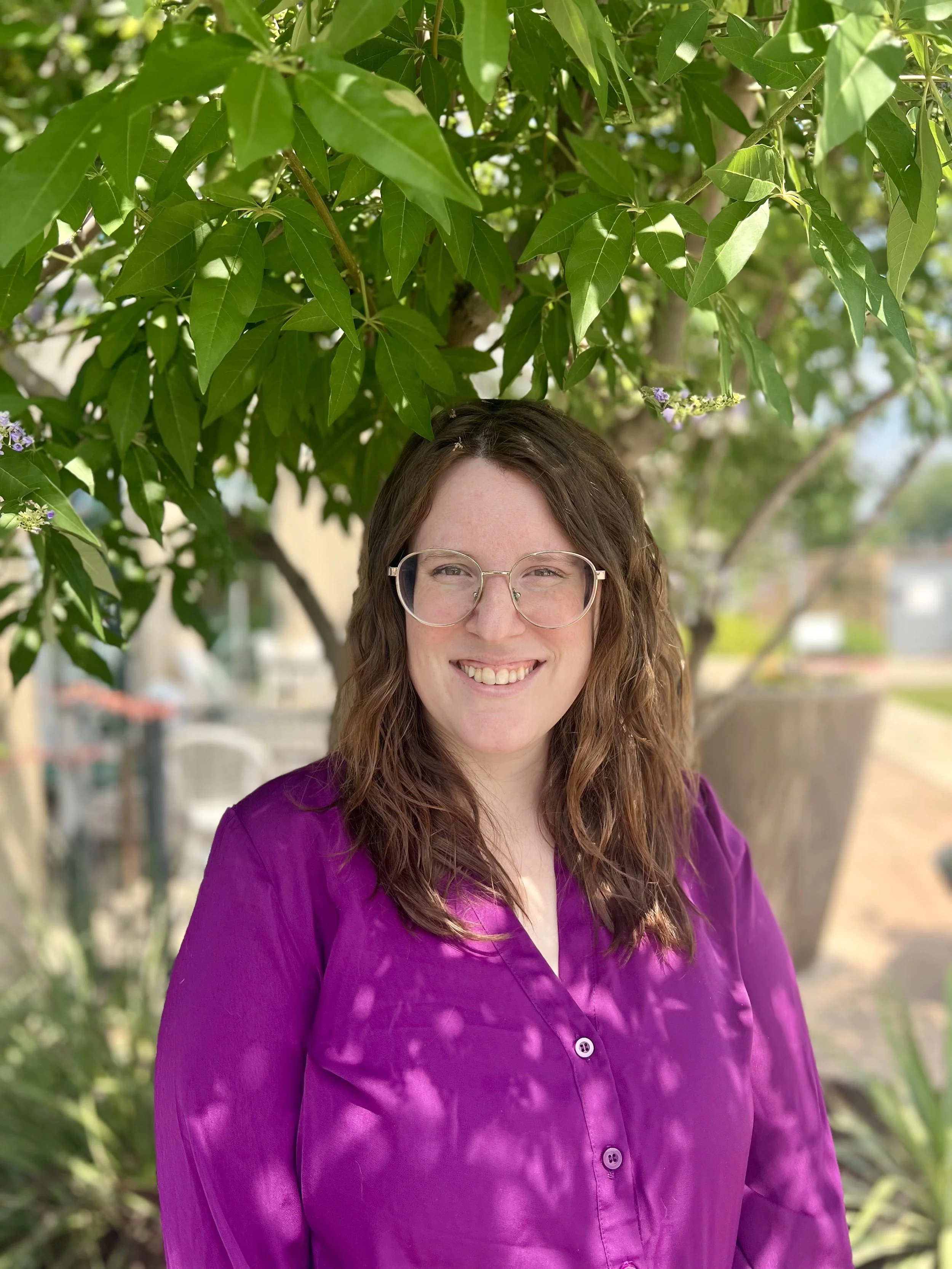 A smiling woman with wavy brown hair and glasses wearing a purple blouse, standing outdoors under a leafy tree.