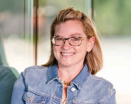 Smiling woman with glasses and shoulder-length brown hair, wearing a denim jacket, indoors with a blurred greenery background.