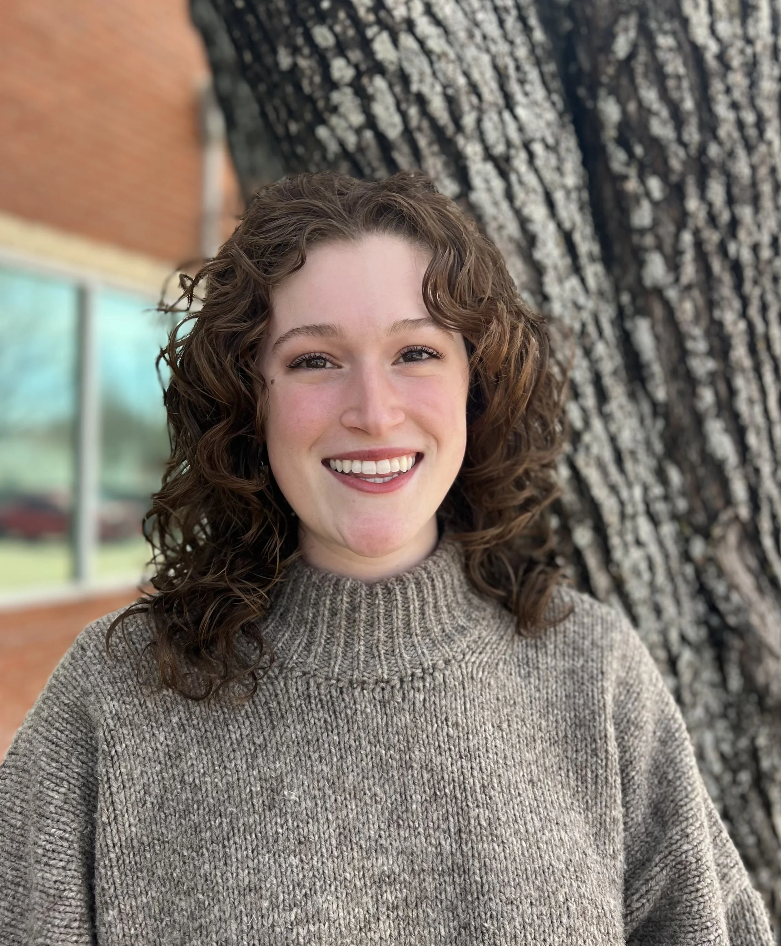 A woman with curly brown hair smiling outdoors near a large tree with textured bark, wearing a beige knit sweater.