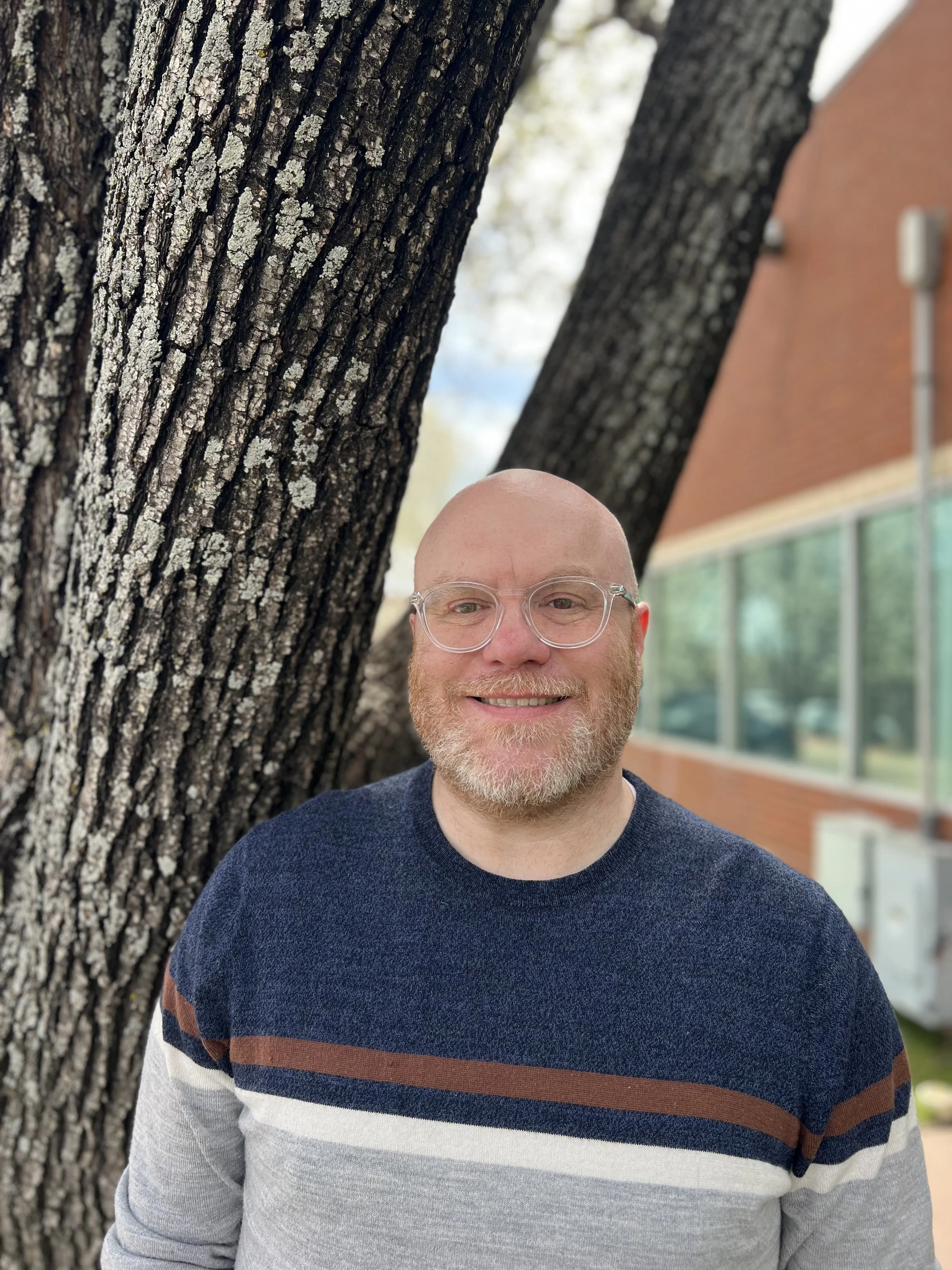 A smiling man with glasses, a beard, and a striped sweater standing outdoors near a large tree with rough bark, with a brick building and windows in the background.