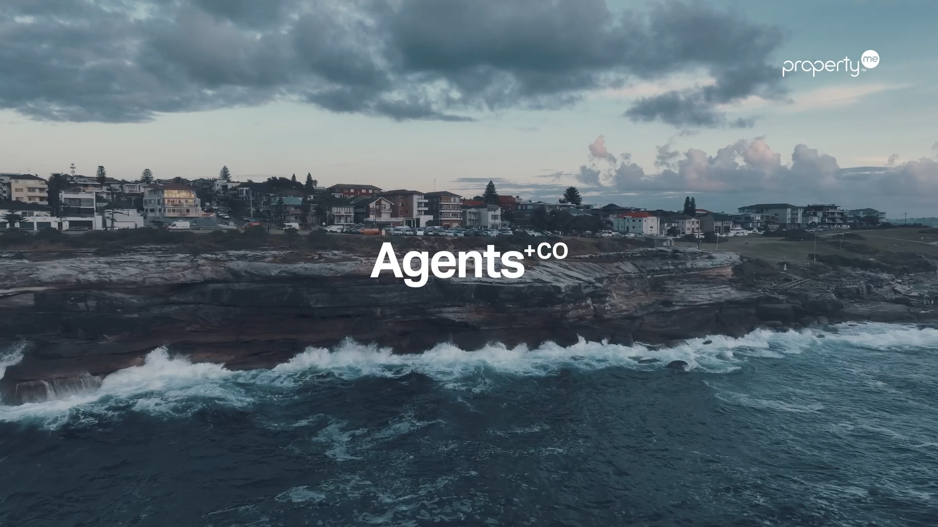 Coastal residential area with houses on cliffs overlooking the ocean under cloudy sky with the text 'Agents+co' in the center and 'property' in the upper right corner.