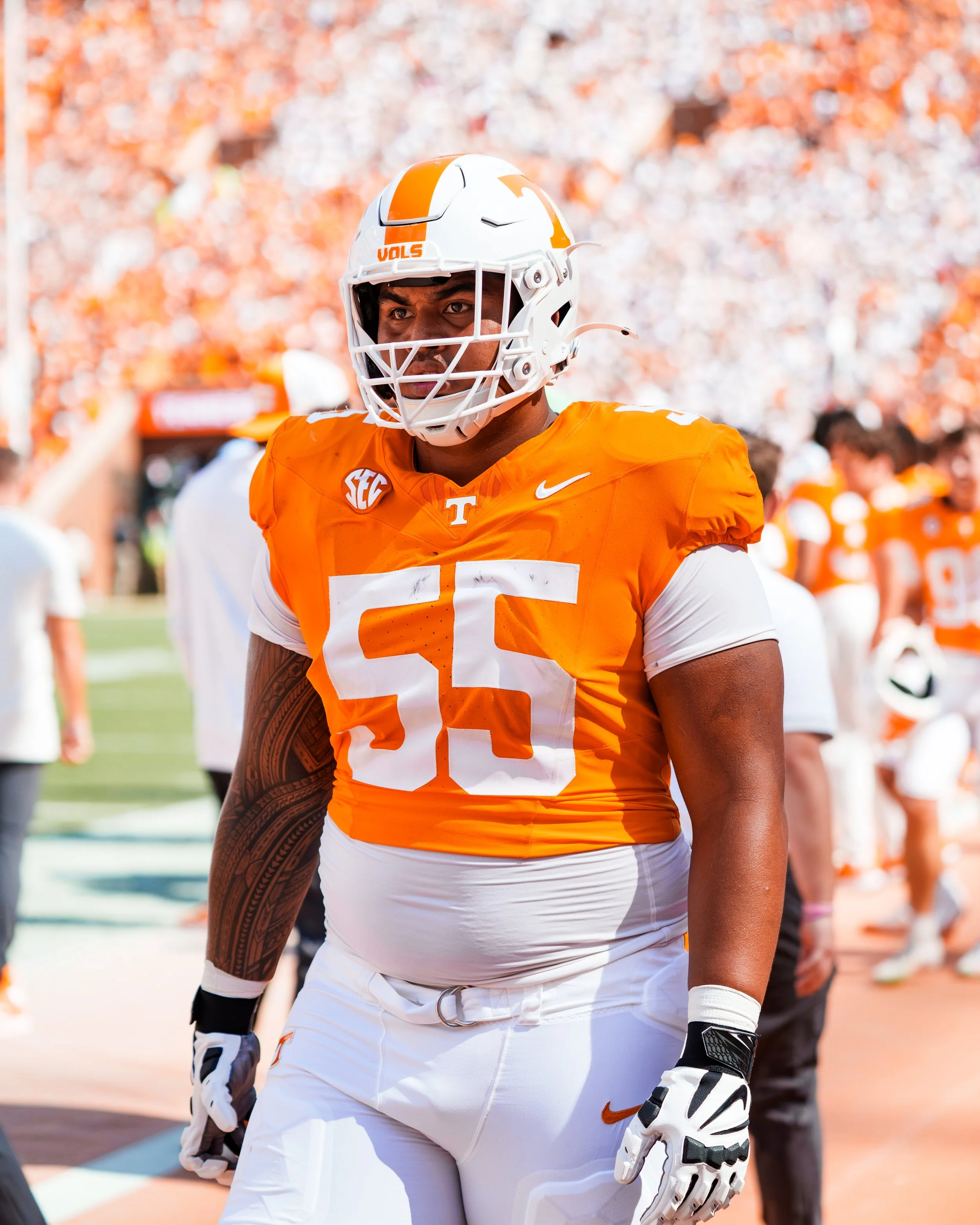 An American football player wearing an orange jersey with the number 55, white pants, a white helmet, and gloves, standing on the field during a game, with fans in the background.