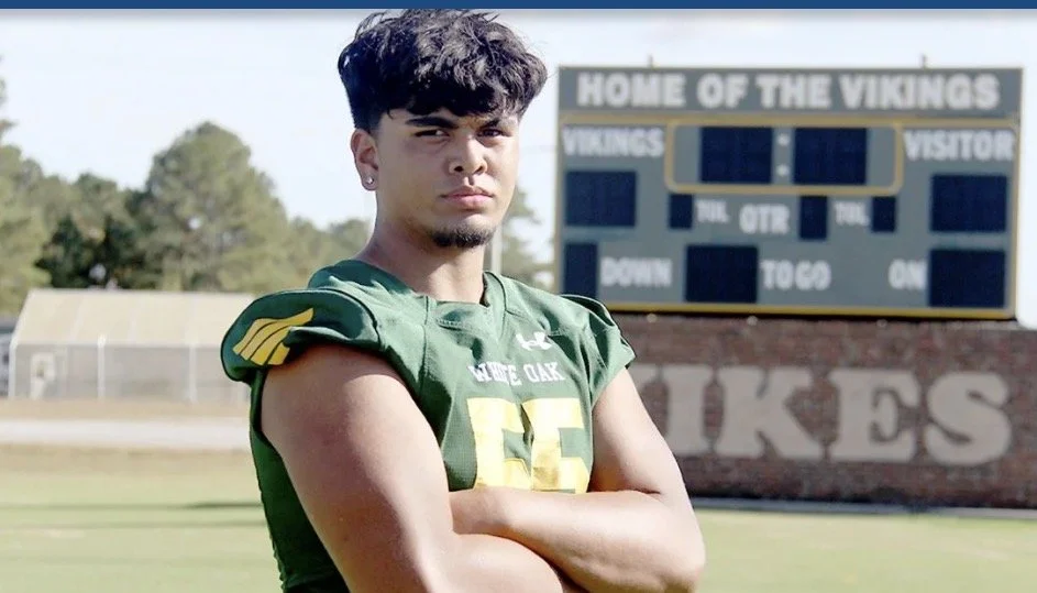Young male football player with crossed arms standing on a football field, scoreboard and school sign in the background.