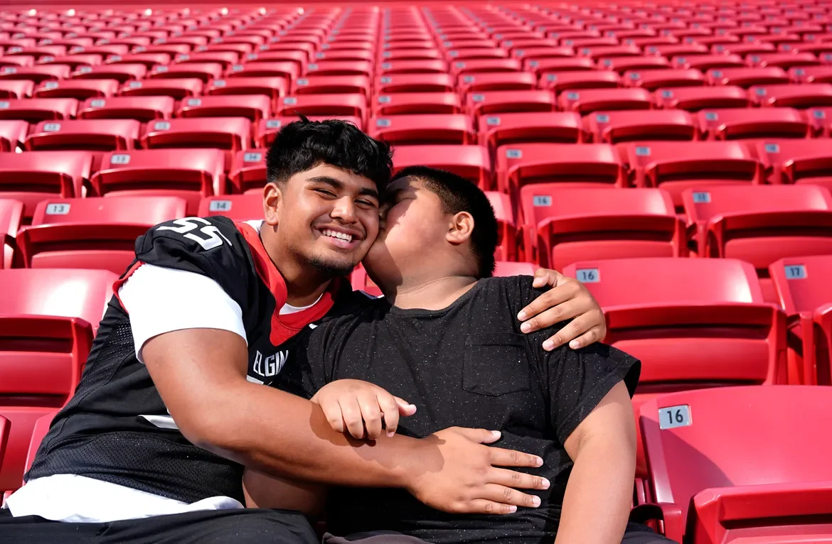 Two men sitting in red stadium seats, one kissing the other's cheek, both smiling, bench marked with seat numbers.