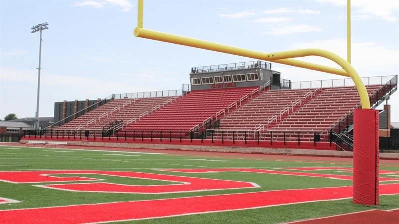 Empty football stadium with red and green turf field, yellow goalpost, and bleachers in the background.