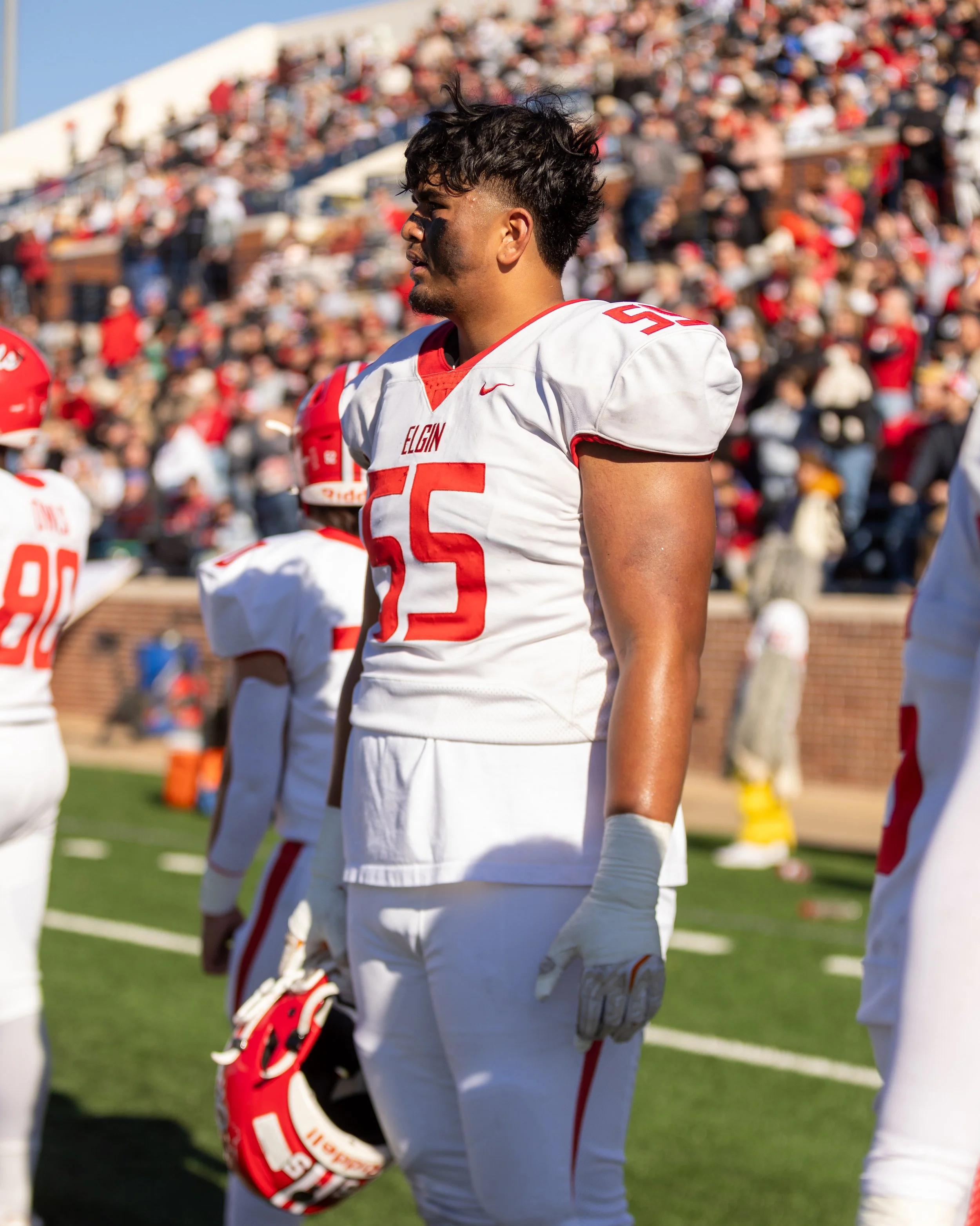 Football player in white uniform with red accents, holding helmet, standing on field with crowd in stands in background.