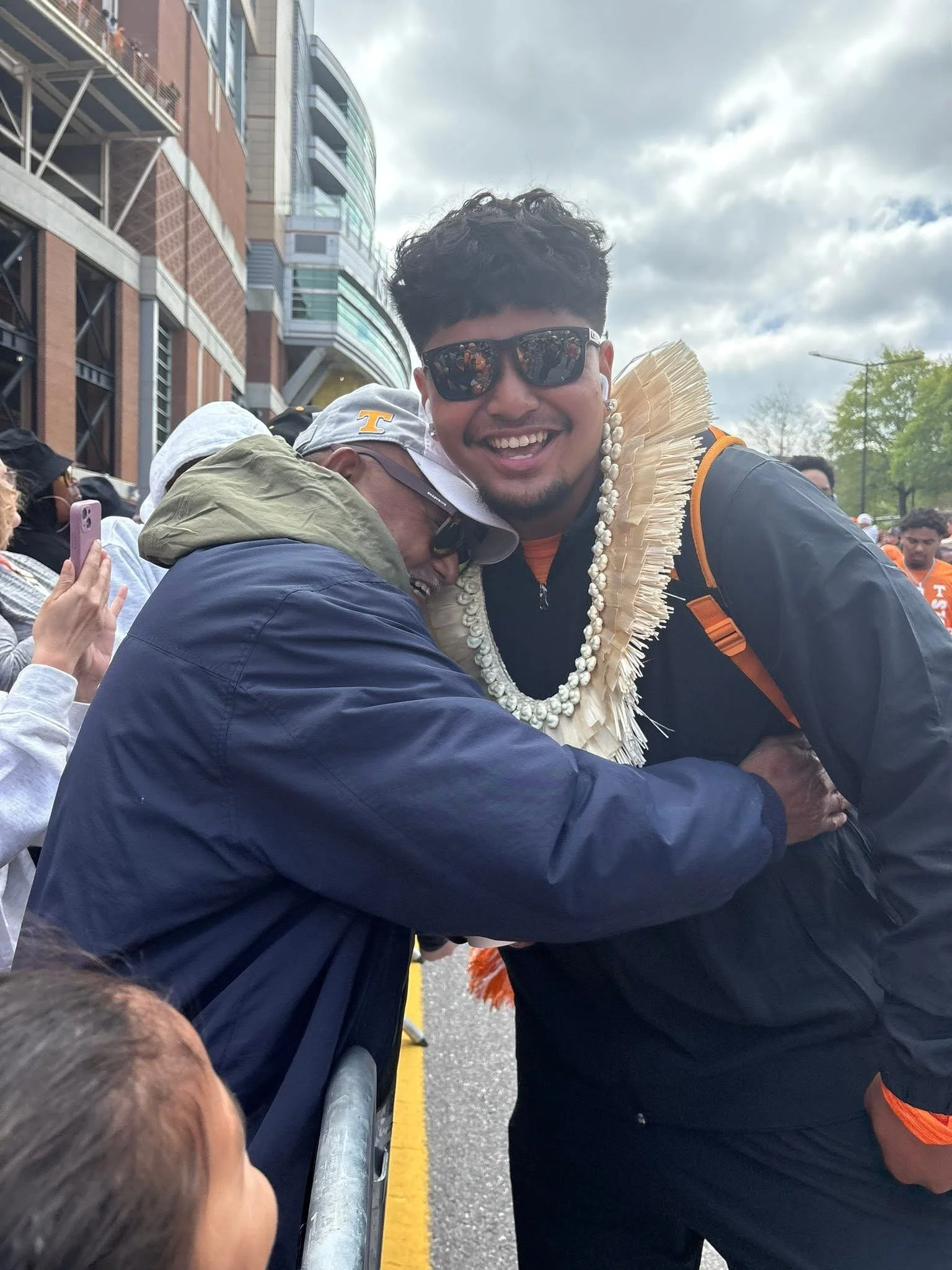 A man wearing sunglasses and a black jacket with orange accents is smiling and hugging someone in front of him at an outdoor event. The man in the front is wearing a traditional Hawaiian lei. There are several other people in the background, some taking photos and wearing casual clothing, and the setting appears to be a city street with modern buildings and cloudy skies.
