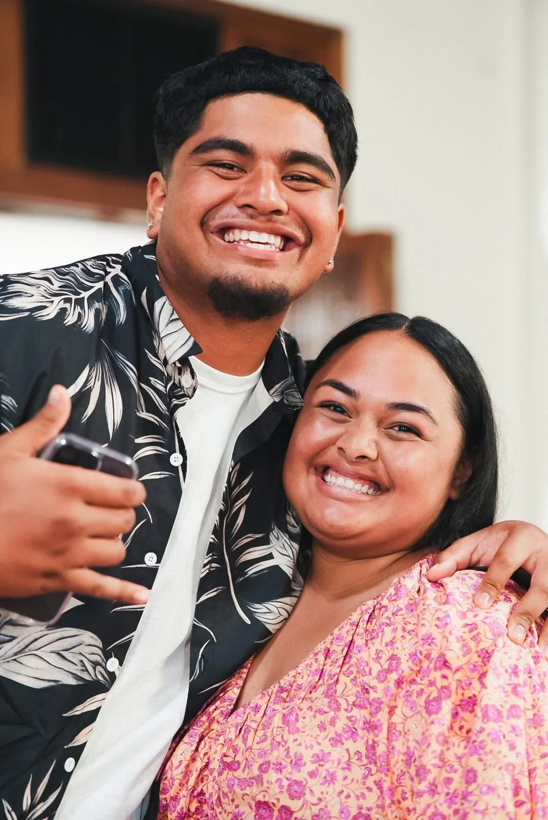 A young man and woman smiling and embracing each other in a friendly pose, with the man making a shaka sign with his hand. The man has short black hair and a goatee, wearing a black and white patterned shirt. The woman has long black hair and is wearing a pink and yellow floral blouse.