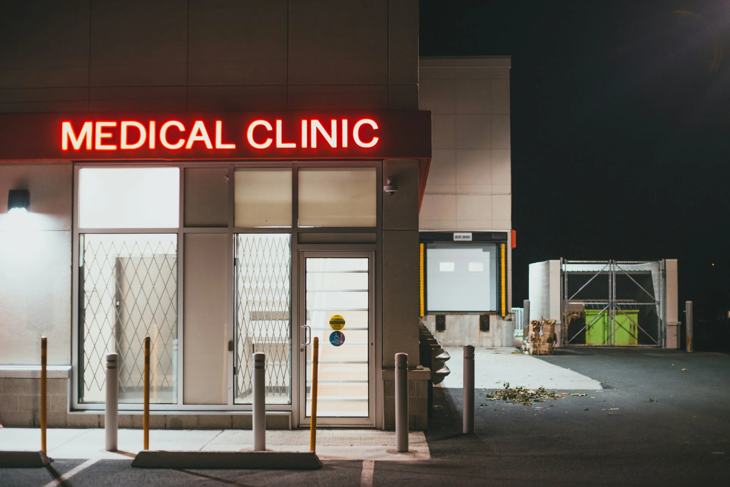 Night view of a medical clinic with a bright red illuminated sign reading 'MEDICAL CLINIC' above the entrance. The entrance has glass doors with security bars, and there is a sidewalk with posts in front of the building. The background shows a parking lot and some dumpsters.