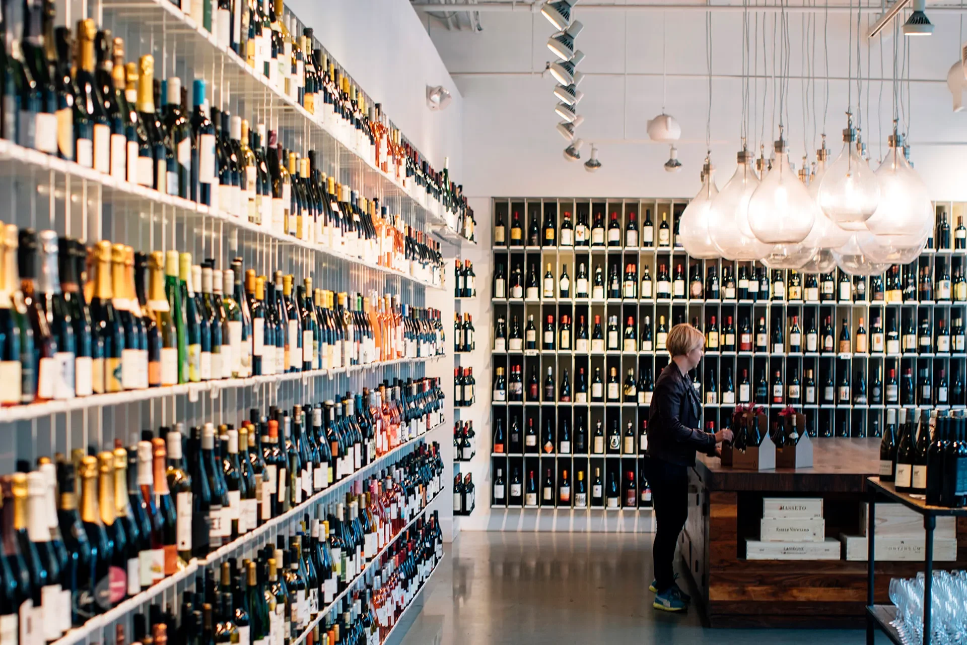 A woman shopping for wine in a modern store with shelves filled with wine bottles, and a large wooden display table, white walls, and unique hanging glass light fixtures.