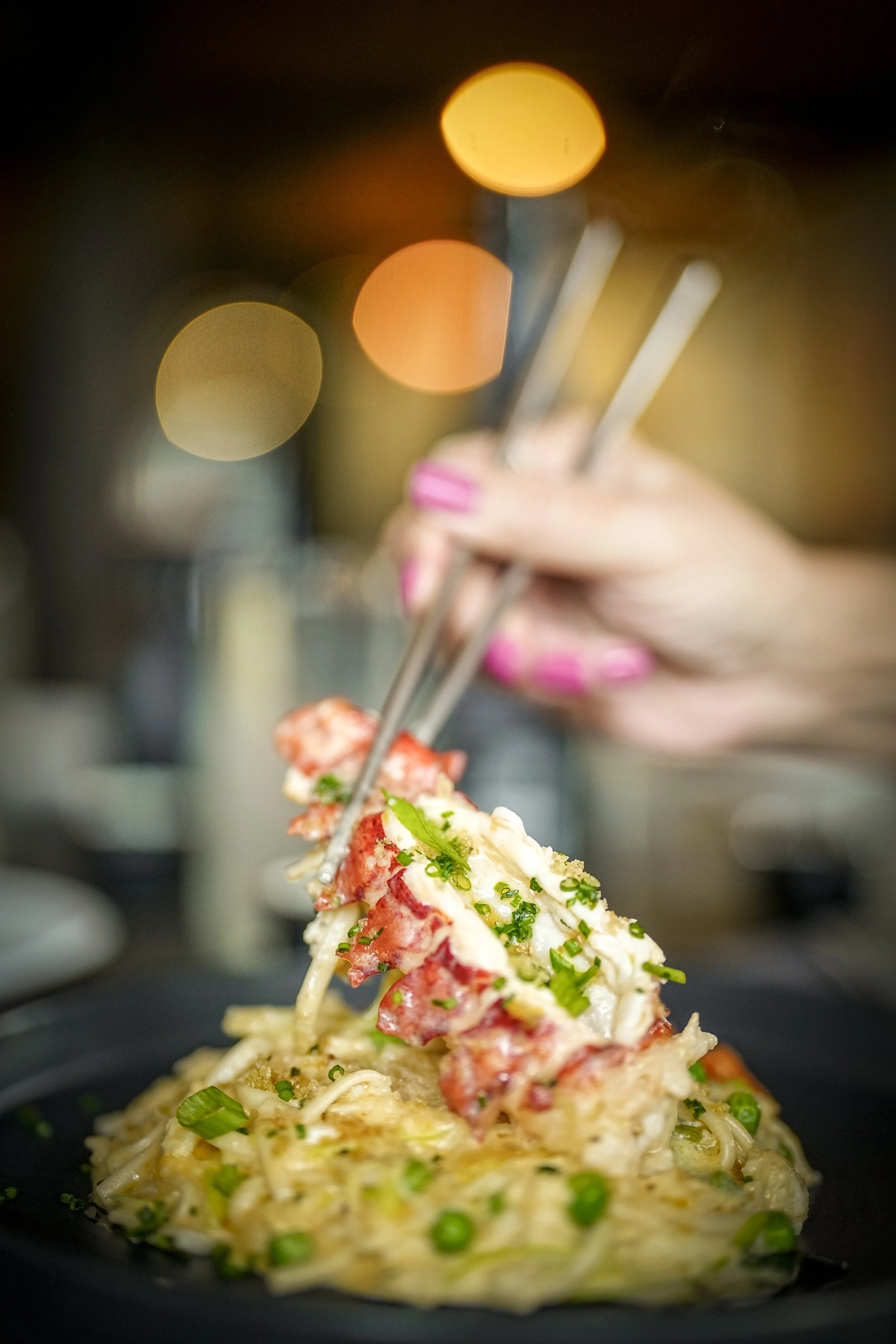 Close-up of a person holding chopsticks with lobster tail and crab leg on a bed of scrambled eggs and vegetables, in a restaurant setting with bokeh lights in the background.