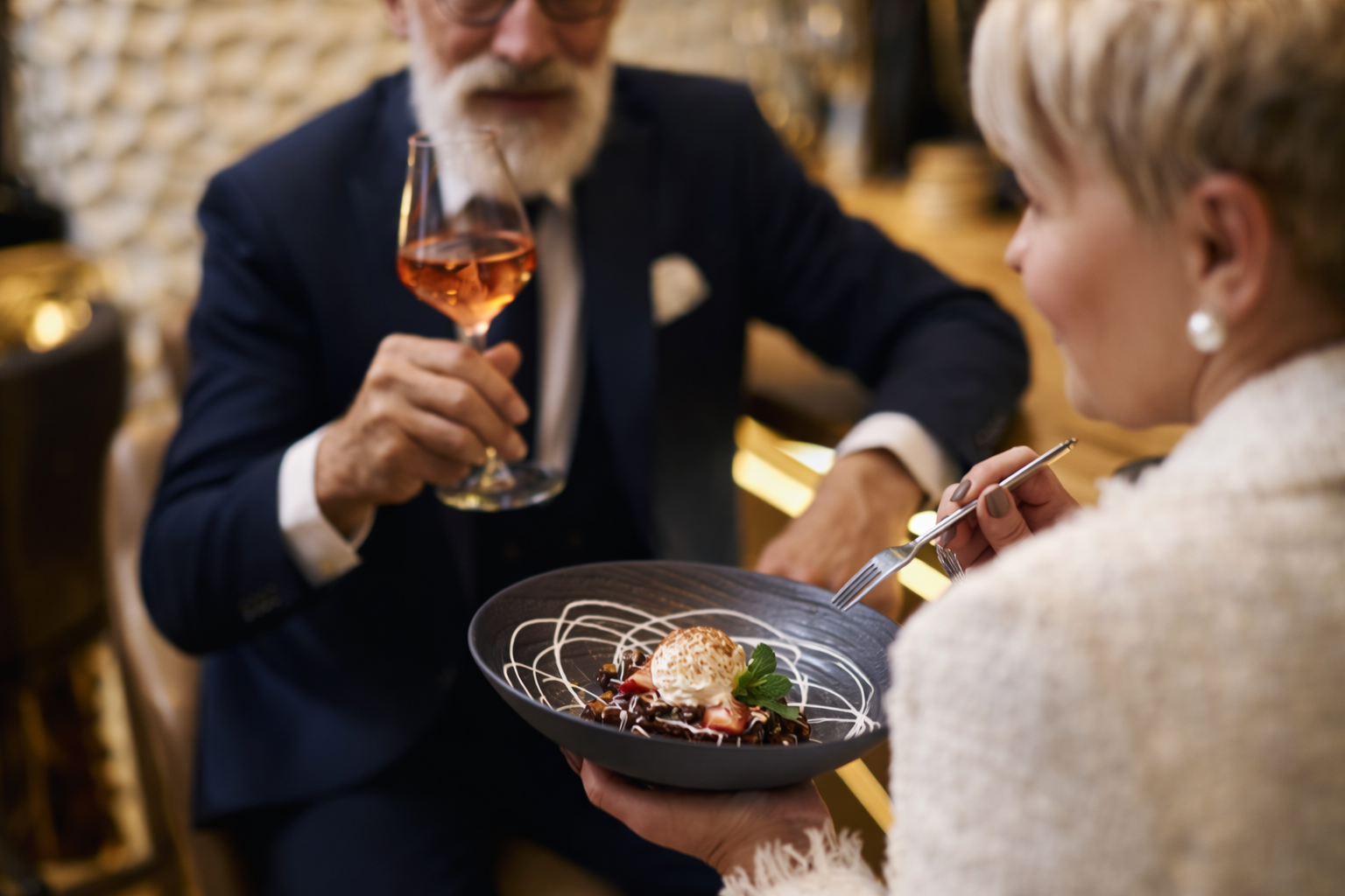 A man in a dark suit holds a glass of rosé wine, smiling at a woman who is holding a fork and a dessert plate with ice cream and chocolate. They sit in a warmly lit, elegant setting.