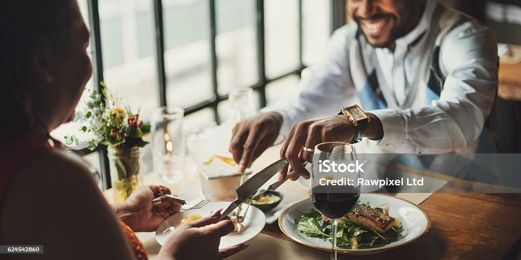 A man and woman dining at a restaurant, with the man serving food to the woman. The man is smiling, dressed in a white shirt with an argyle pattern, and is wearing a watch and ring. The woman is holding a fork. There is a glass of red wine on the table, along with a plate of steak and greens, and a small plant in the background.