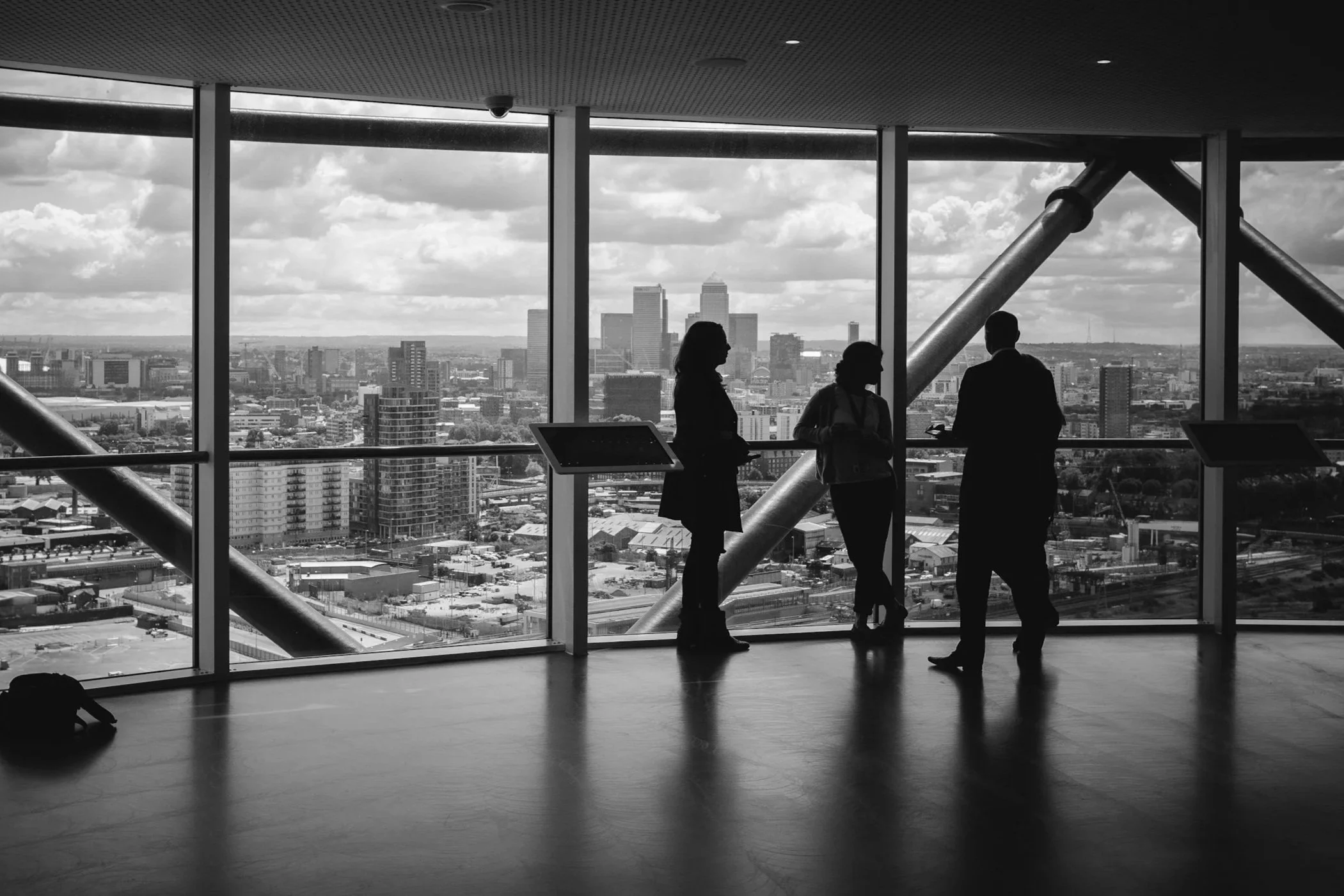 Black and white image of three silhouetted people standing inside a modern building, looking out through large glass windows at a city skyline with structural beams.
