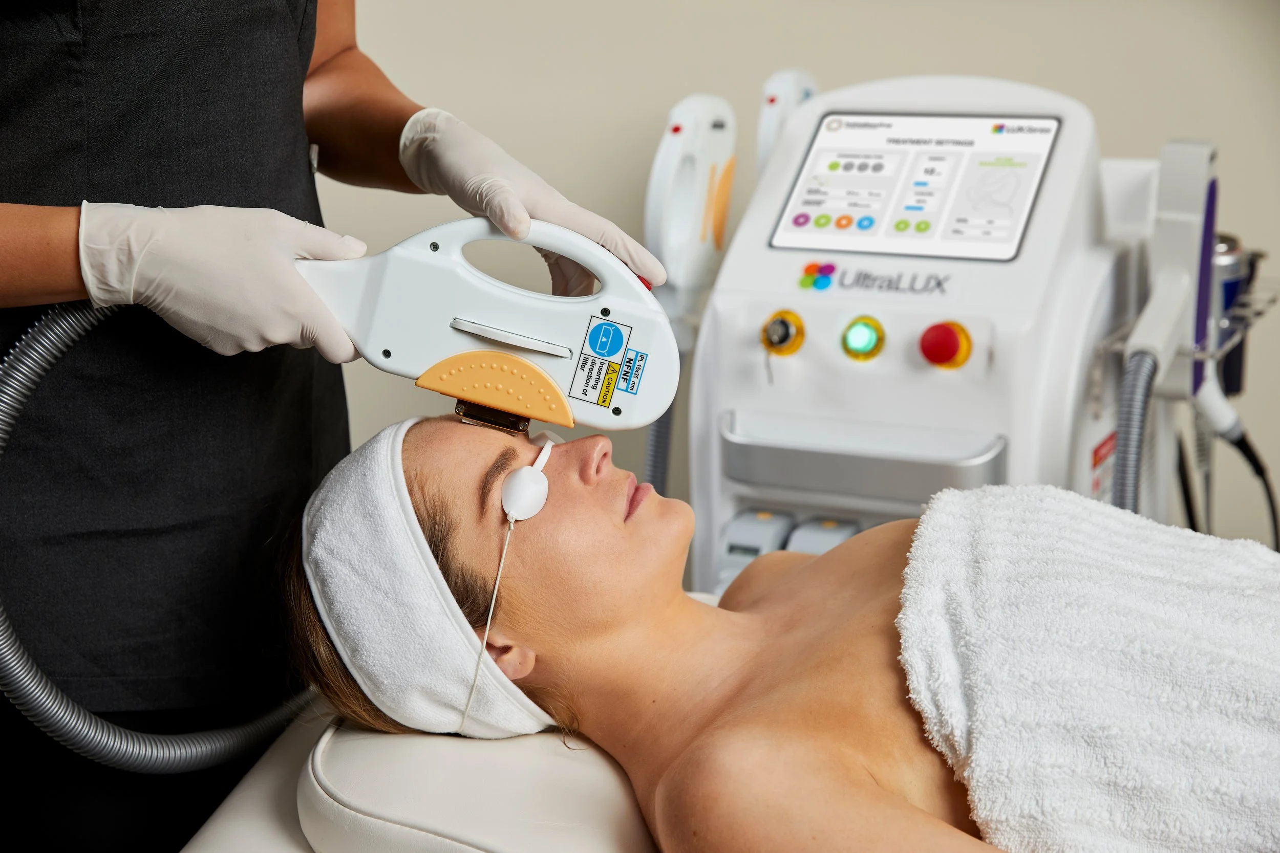 A woman lying on a medical bed with her eyes closed, receiving a laser treatment on her face from a healthcare professional holding a laser device. The woman wears a towel over her chest and a headband, and the professional wears gloves. Medical equipment and a control screen are visible in the background.