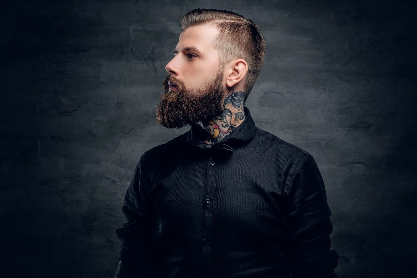 Side profile of a man with a textured beard, slicked-back hair, and tattoos on his neck, wearing a black shirt against a dark gray background.