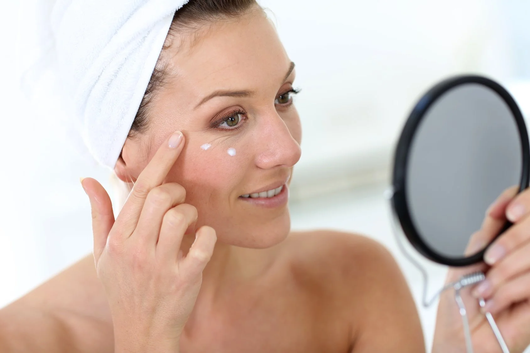 A woman with a white towel on her head applying skincare to her face while looking into a handheld mirror.