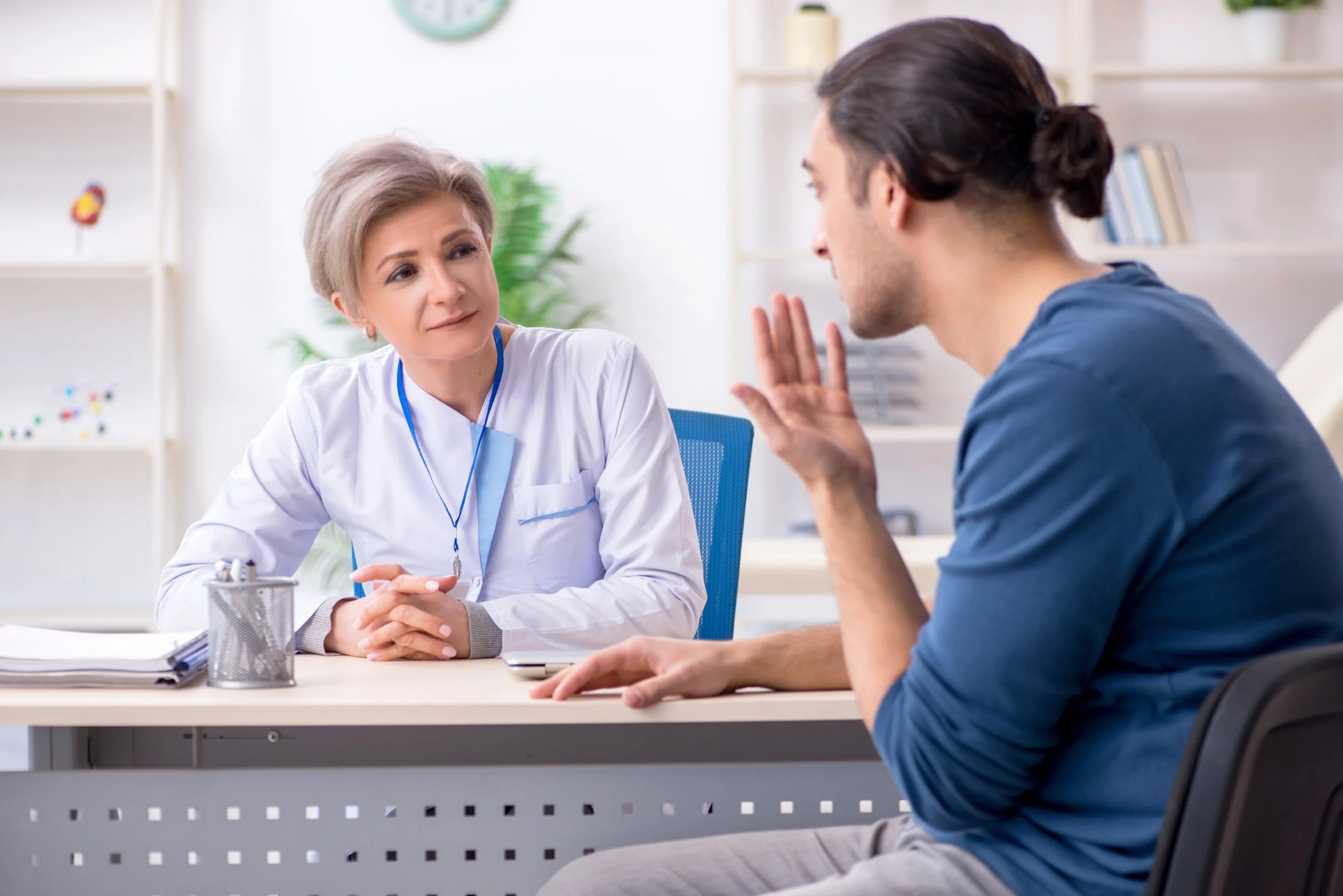 A doctor and a patient having a consultation in a medical office. The doctor, an older woman with short gray hair, is listening attentively, while the young male patient, with long dark hair, is explaining something and gesturing with his hand.