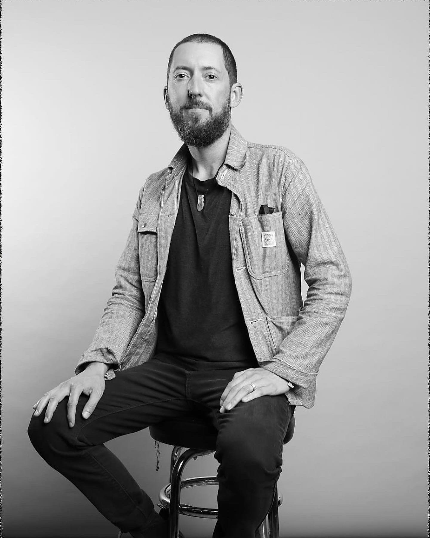 A man with a beard and short hair, sitting on a stool against a plain background, wearing a casual jacket over a dark shirt, with a ring on his finger and some pen in his jacket pocket, looking at the camera.