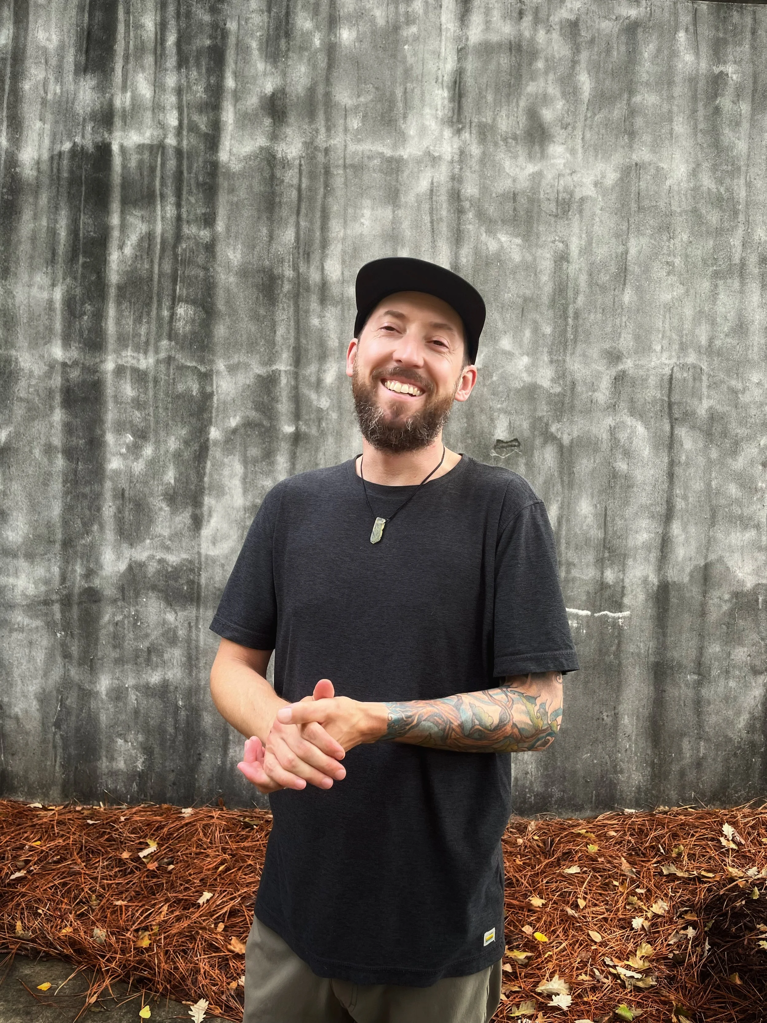 A smiling man with a beard wearing a black T-shirt, a black cap, and a necklace, standing outdoors in front of a textured gray wall and fallen autumn leaves.