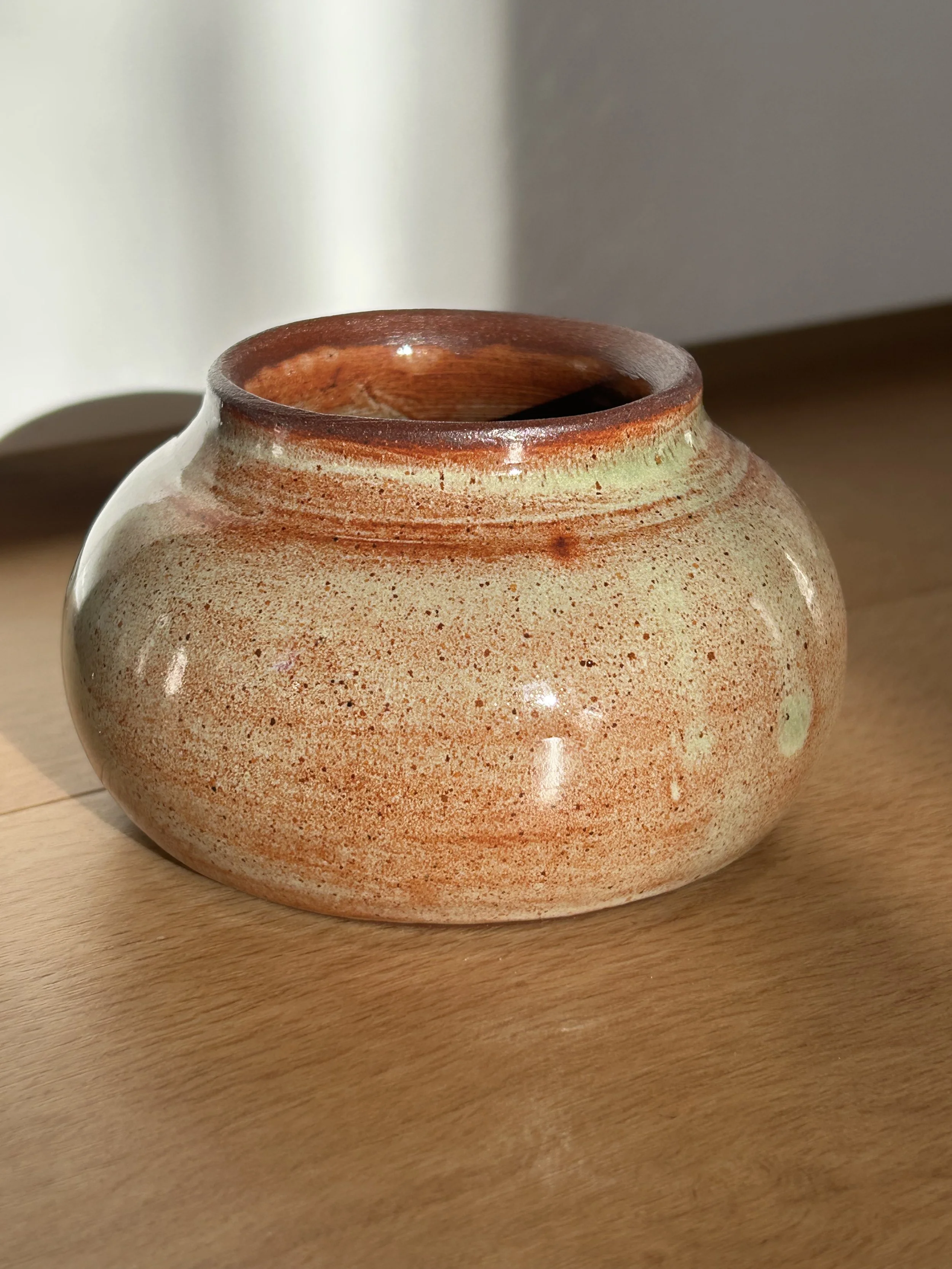 A round ceramic vase with a speckled beige and orange glaze, sitting on a wooden surface with a shadow cast beside it.