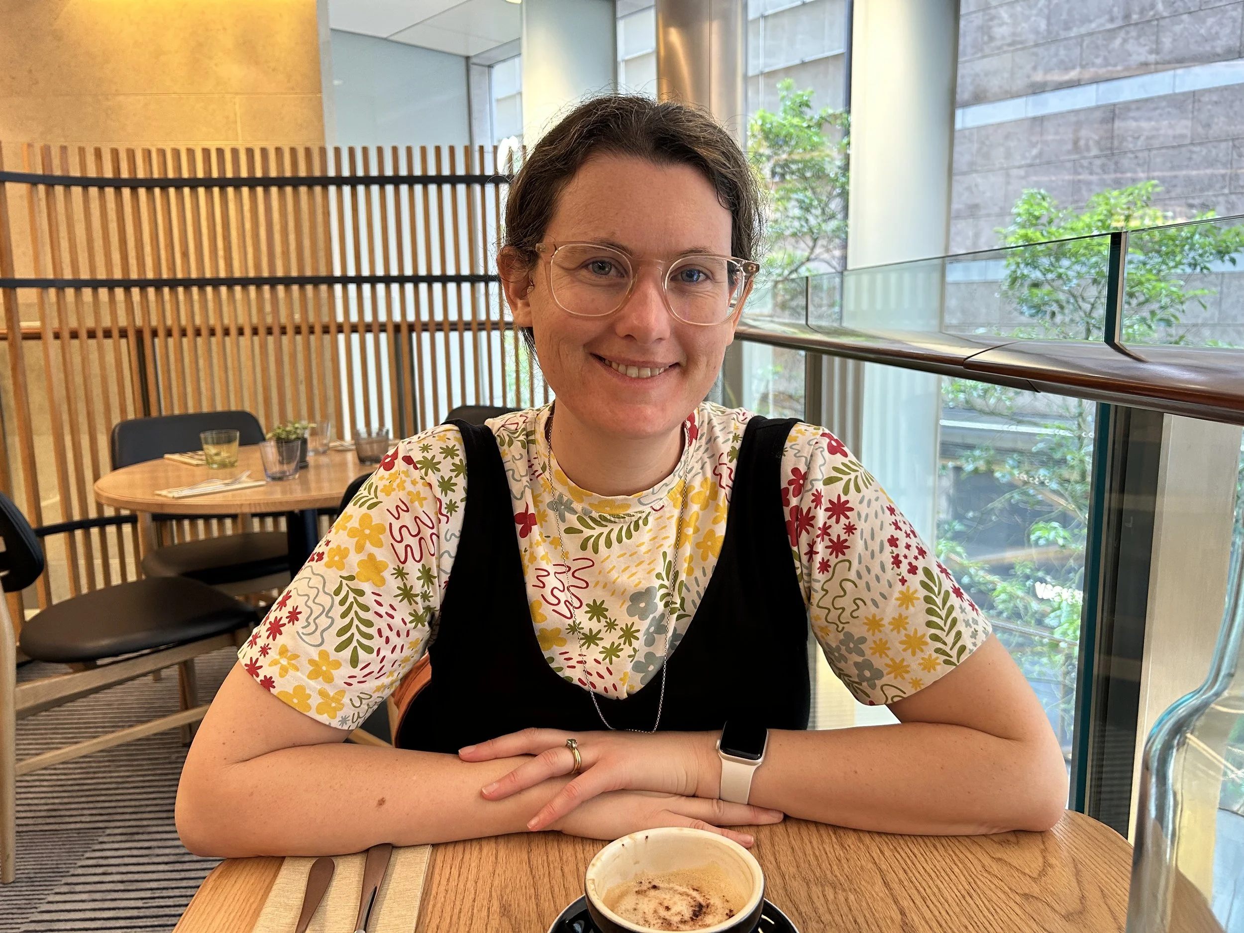 A woman sitting at a wooden table in a modern restaurant with large windows and indoor plants, smiling at the camera, with a cappuccino in front of her.