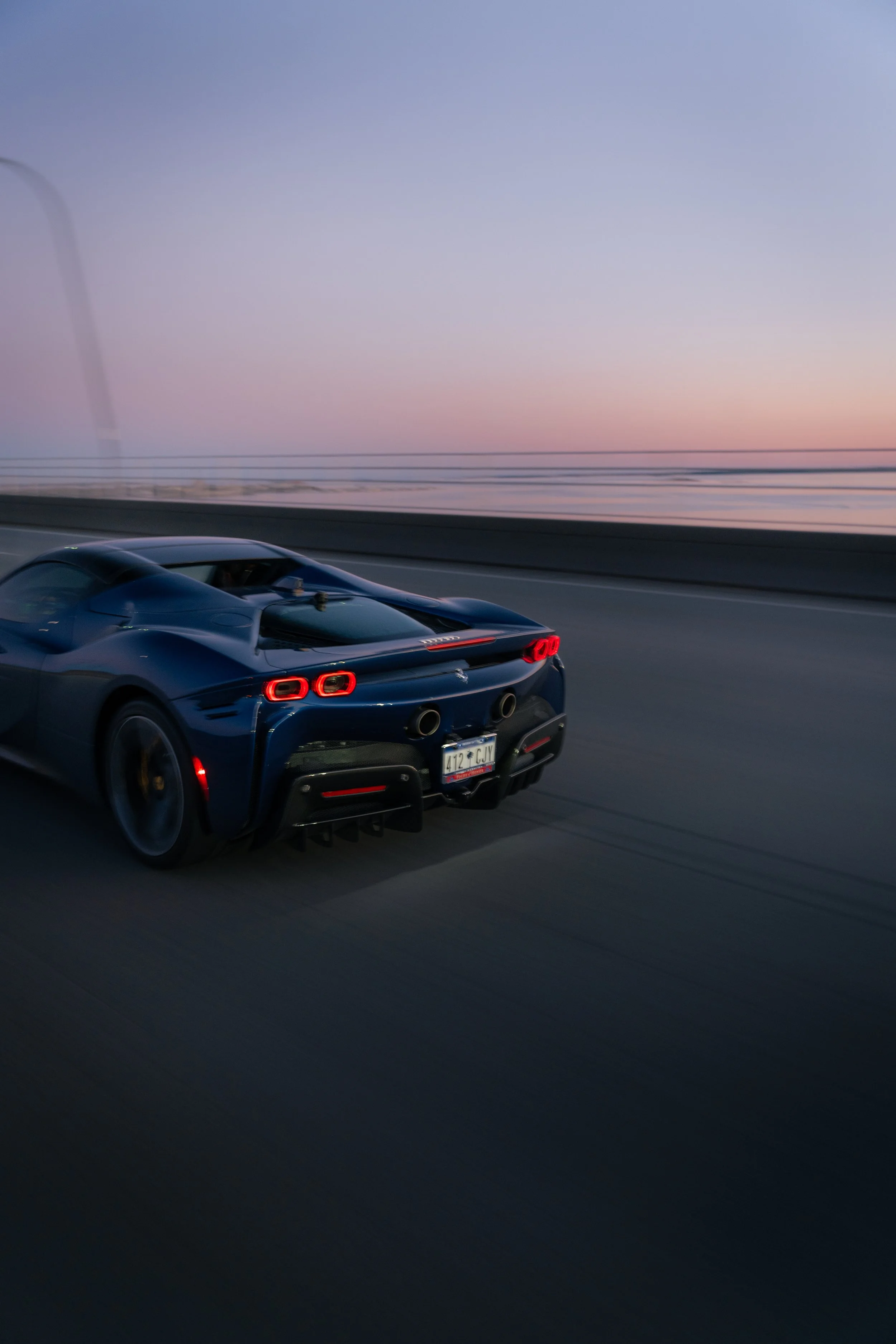 A sleek blue race car driving on a highway at dusk, with a distant landscape and a pastel sky in the background.