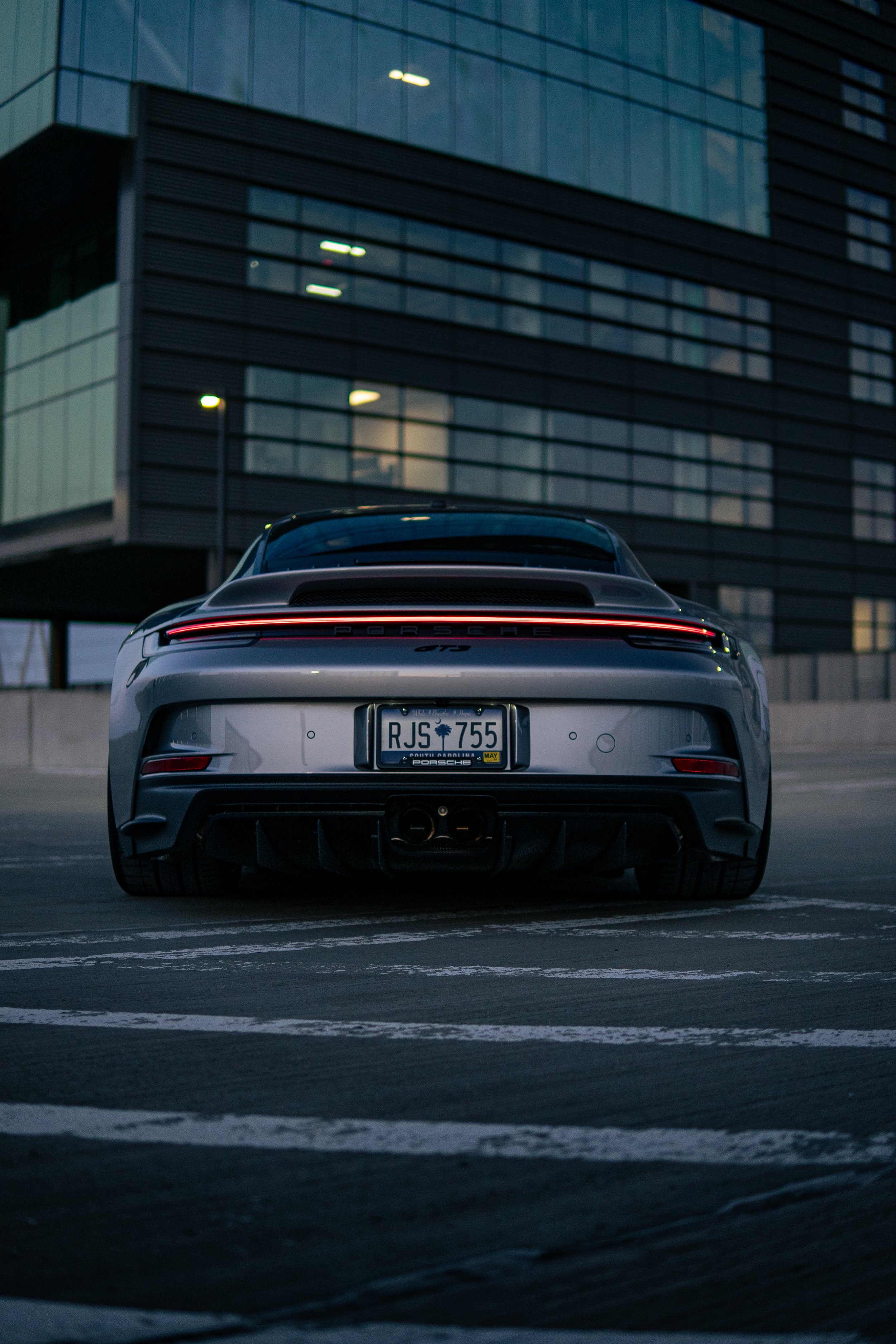 Rear view of a silver Porsche GT3 sports car parked on a rooftop parking lot near a modern glass building during dusk.