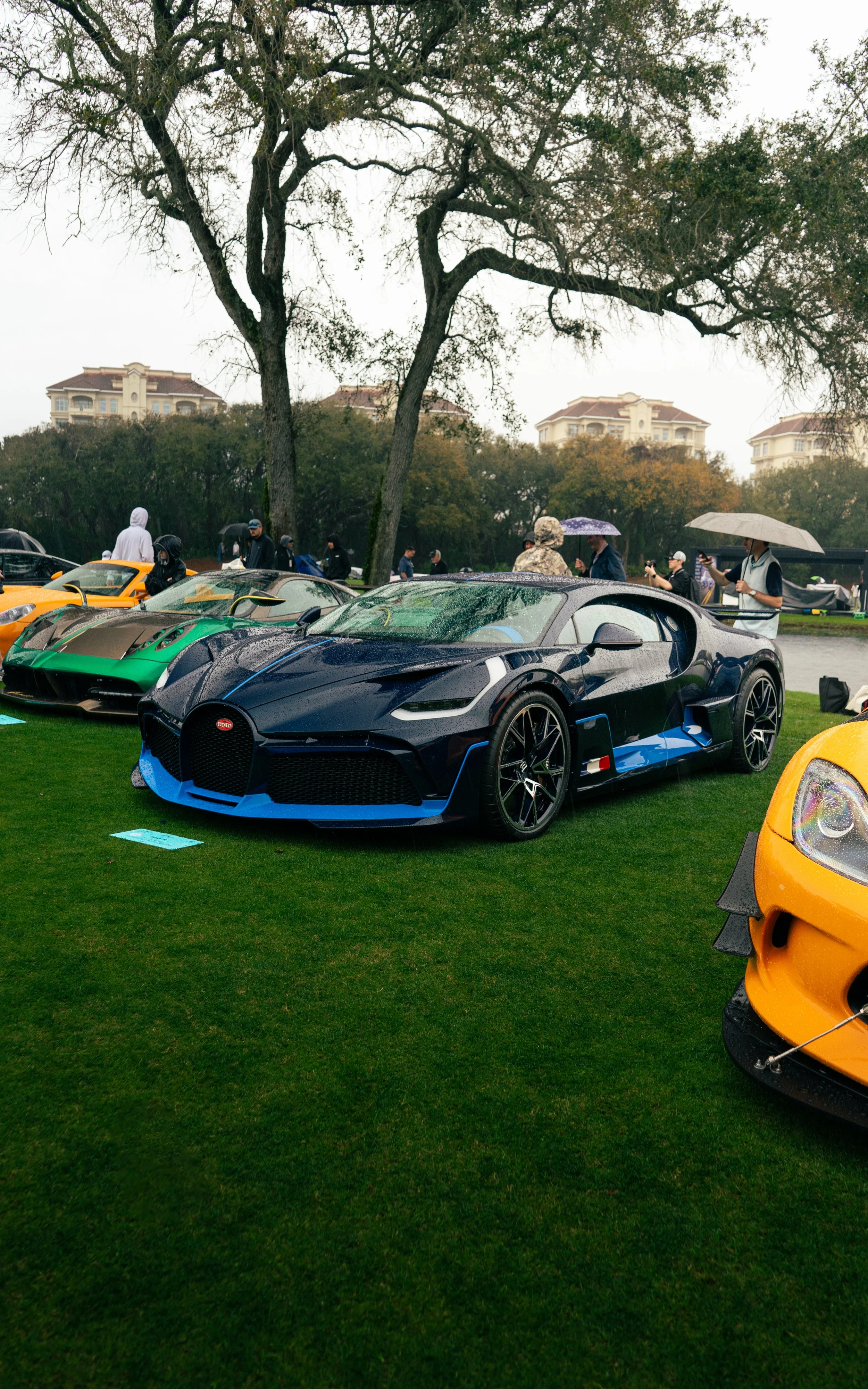 A black and blue luxury sports car on display at a car show, with other exotic cars and people with umbrellas in the background under cloudy weather.