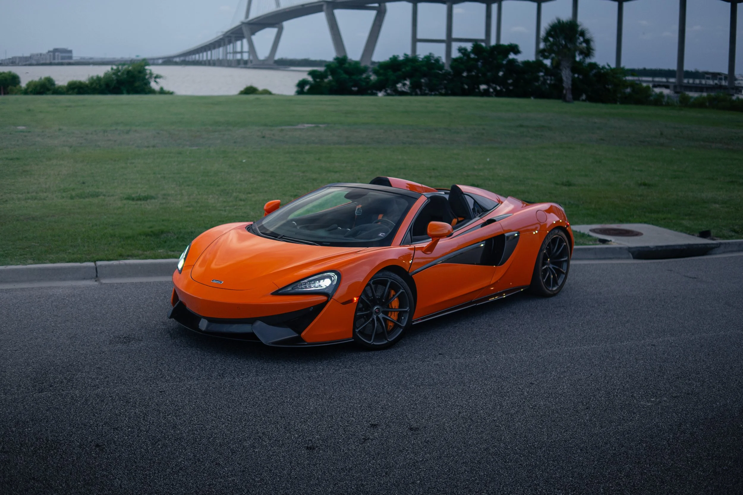Orange sports car parked on a road near a grassy area with a bridge in the background.