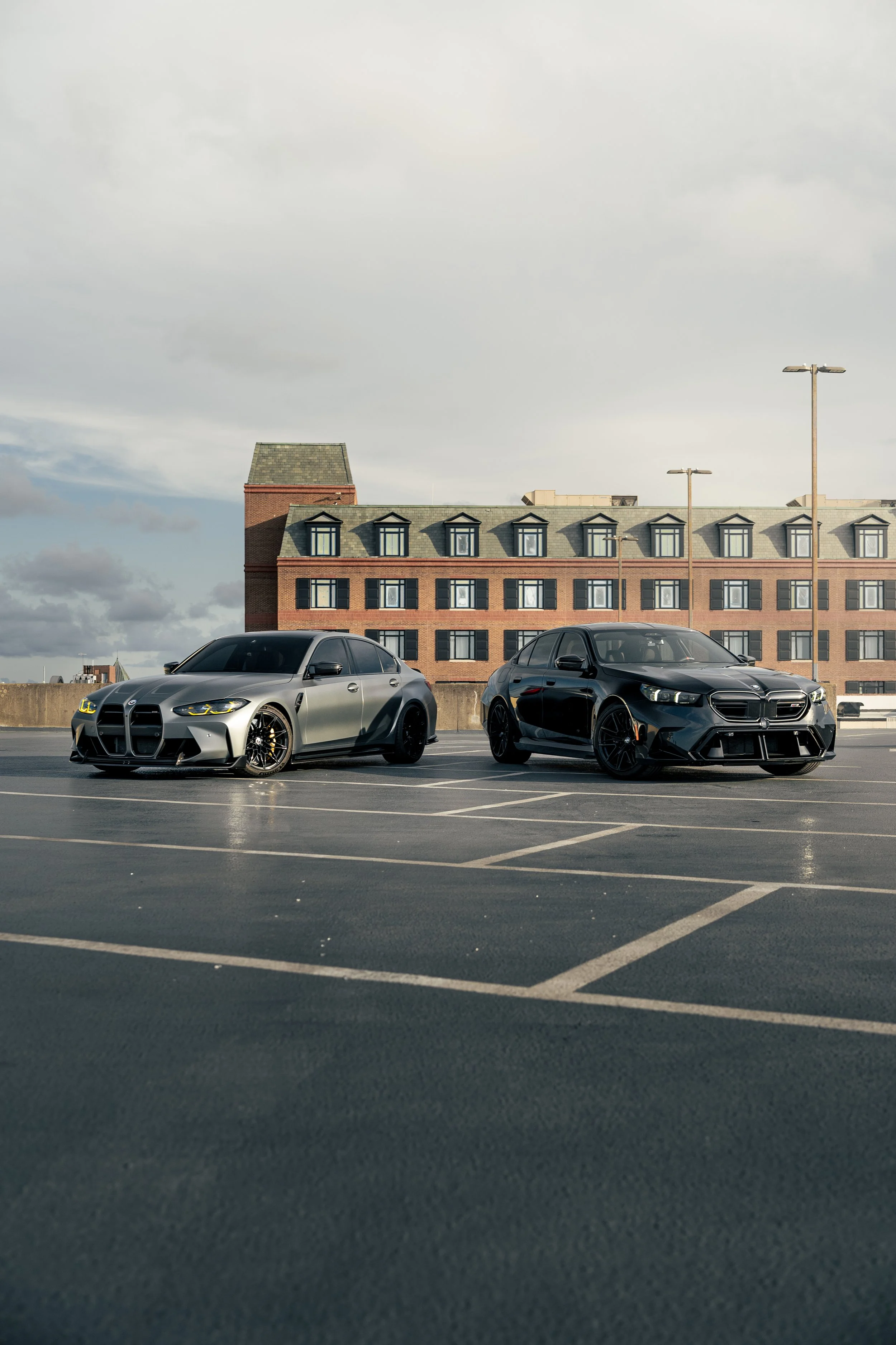 Two modern cars parked in a rooftop parking lot in front of a multi-story brick building under a cloudy sky.