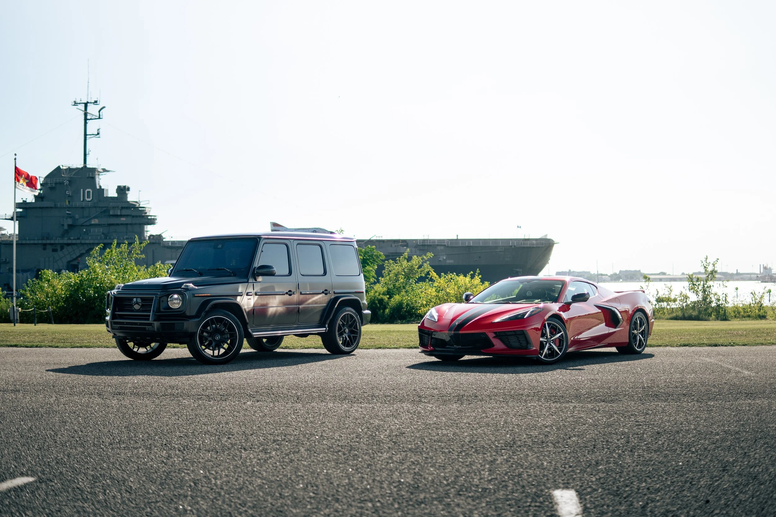 A black Mercedes G-Class SUV and a red sports car parked on a paved surface near grass, with an aircraft carrier docked in the water and a navy flag in the background.