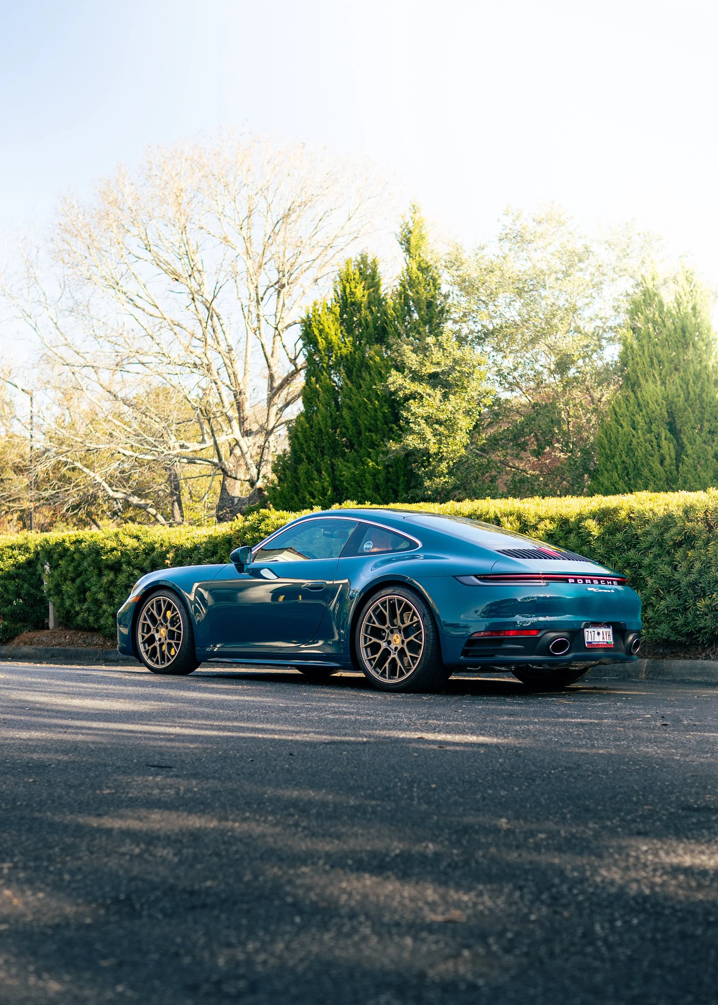 Dark green Porsche sports car parked on the street with trees and bushes in the background on a sunny day.