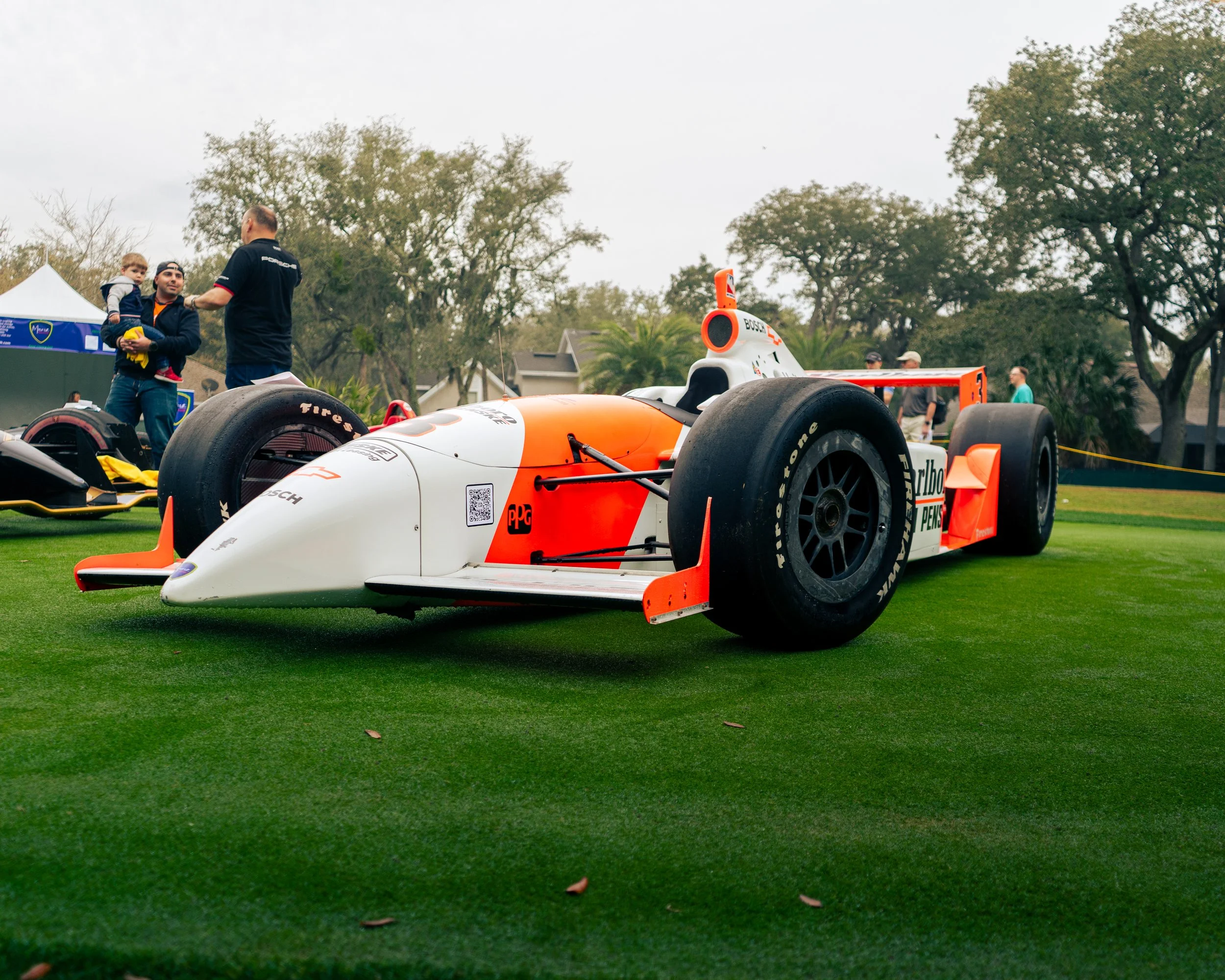 A vintage racing car displayed on a grassy area with people around, including a man holding a child. Trees and buildings are in the background.