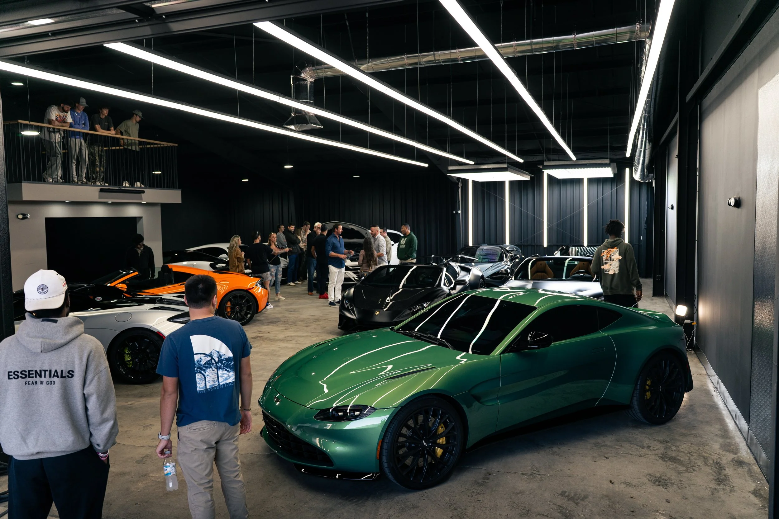Indoor car showroom featuring luxury and sports cars, with people viewing and socializing, illuminated by modern lighting fixtures.
