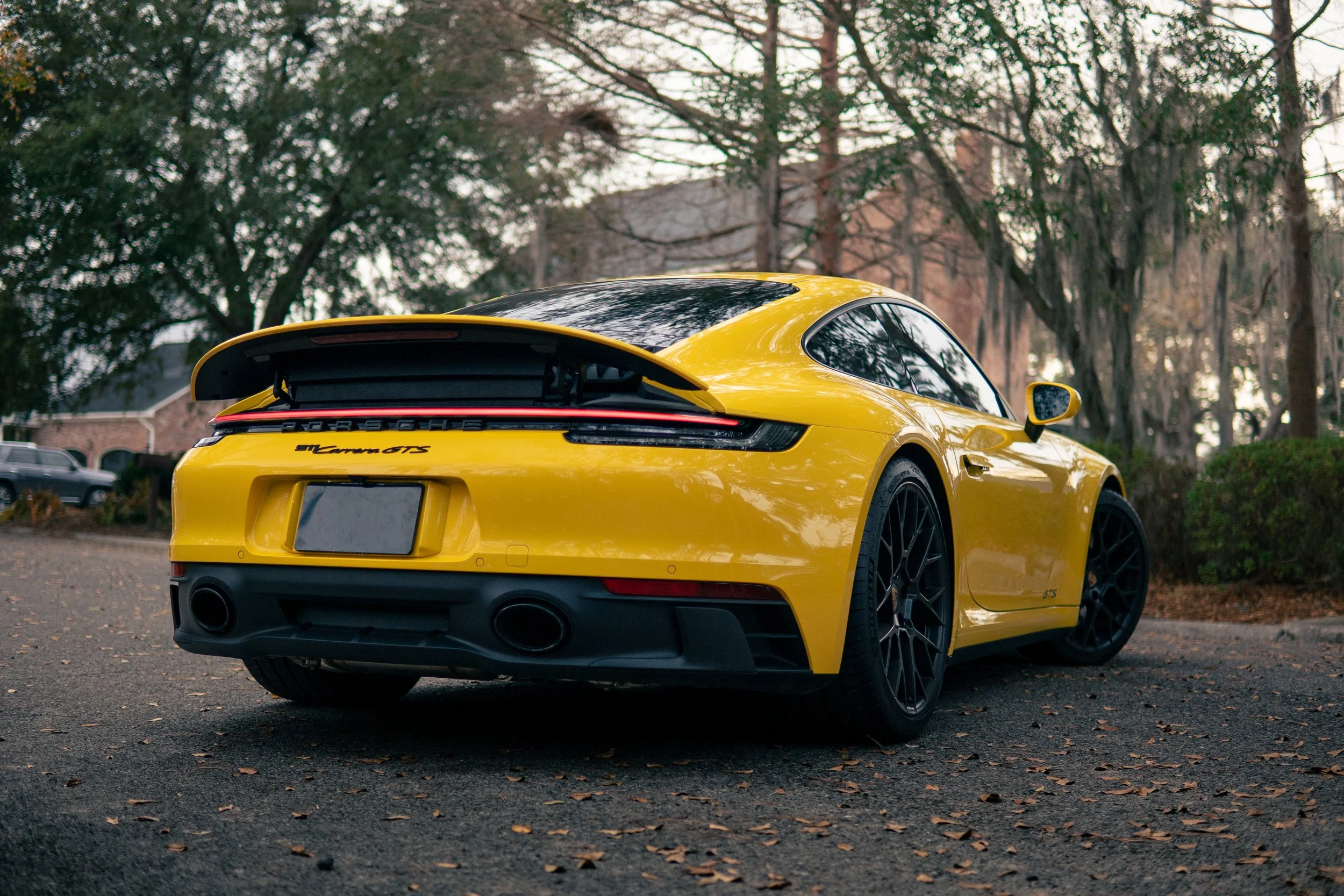 Yellow Porsche 911 Carrera GTS parked on a paved surface with fallen leaves, surrounded by trees and bushes, with a building and other vehicles in the background.