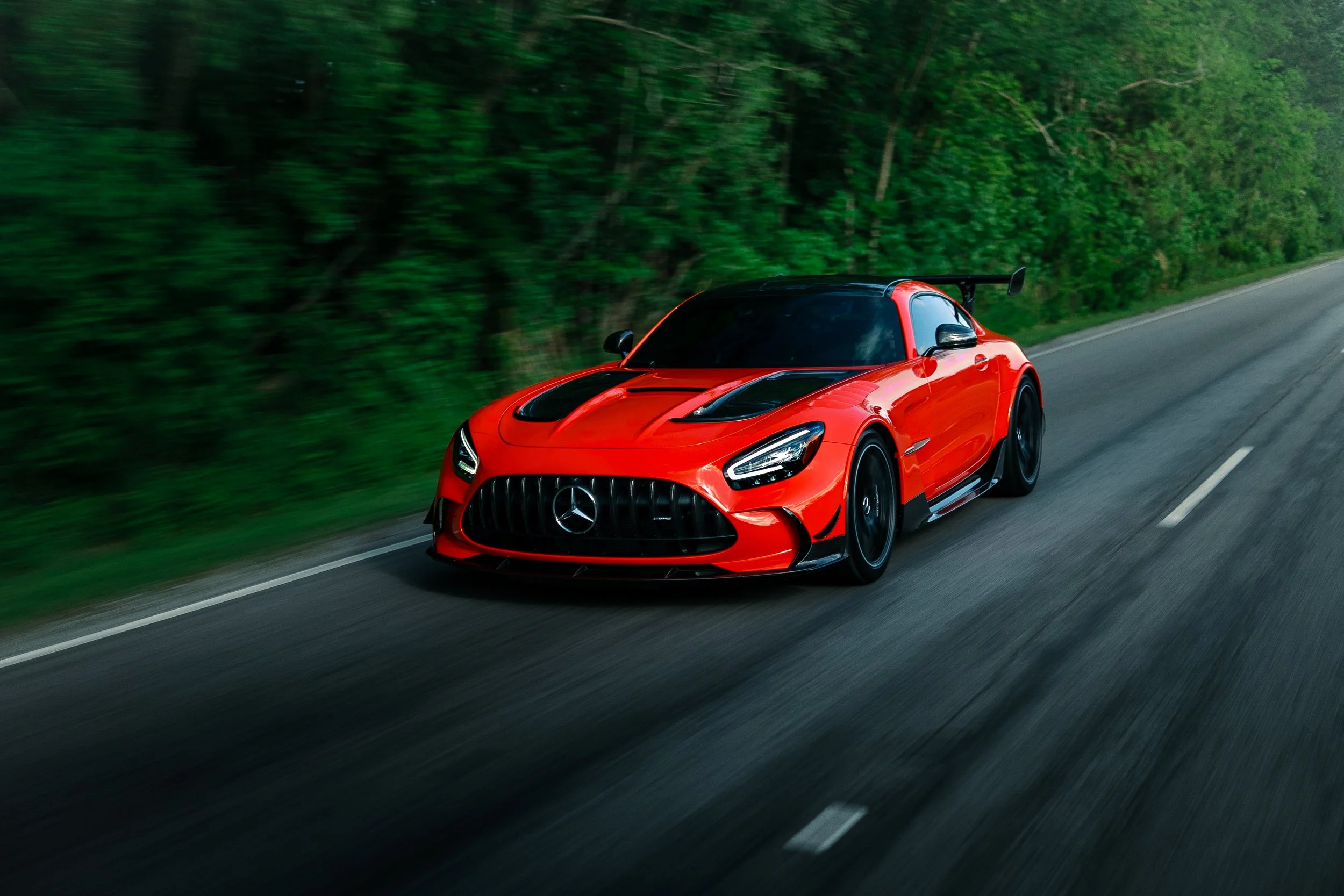 Red Mercedes-Benz race car driving on a two-lane road through a wooded area.