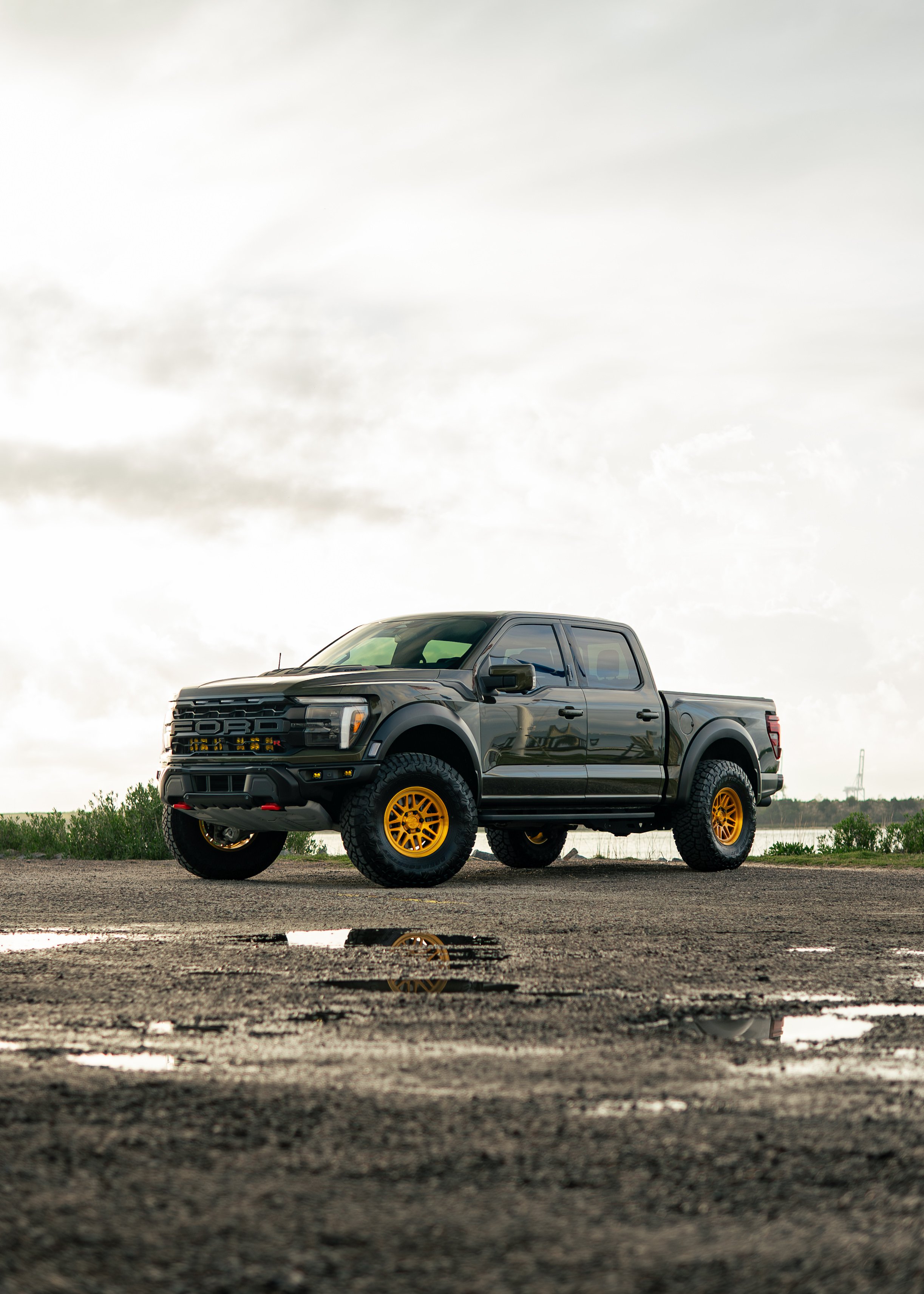 A black Ford F-150 Raptor pickup truck with yellow wheels parked on a gravel surface near a body of water under cloudy skies.