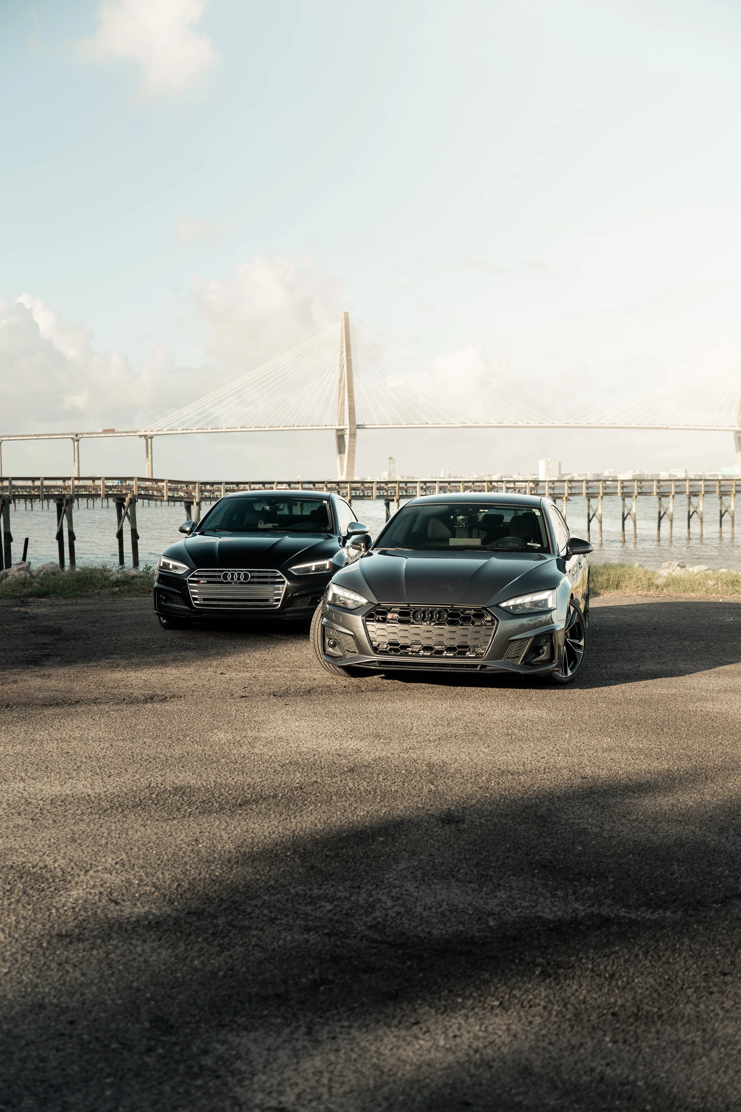 Two black Audi cars parked near a body of water with a bridge in the background.