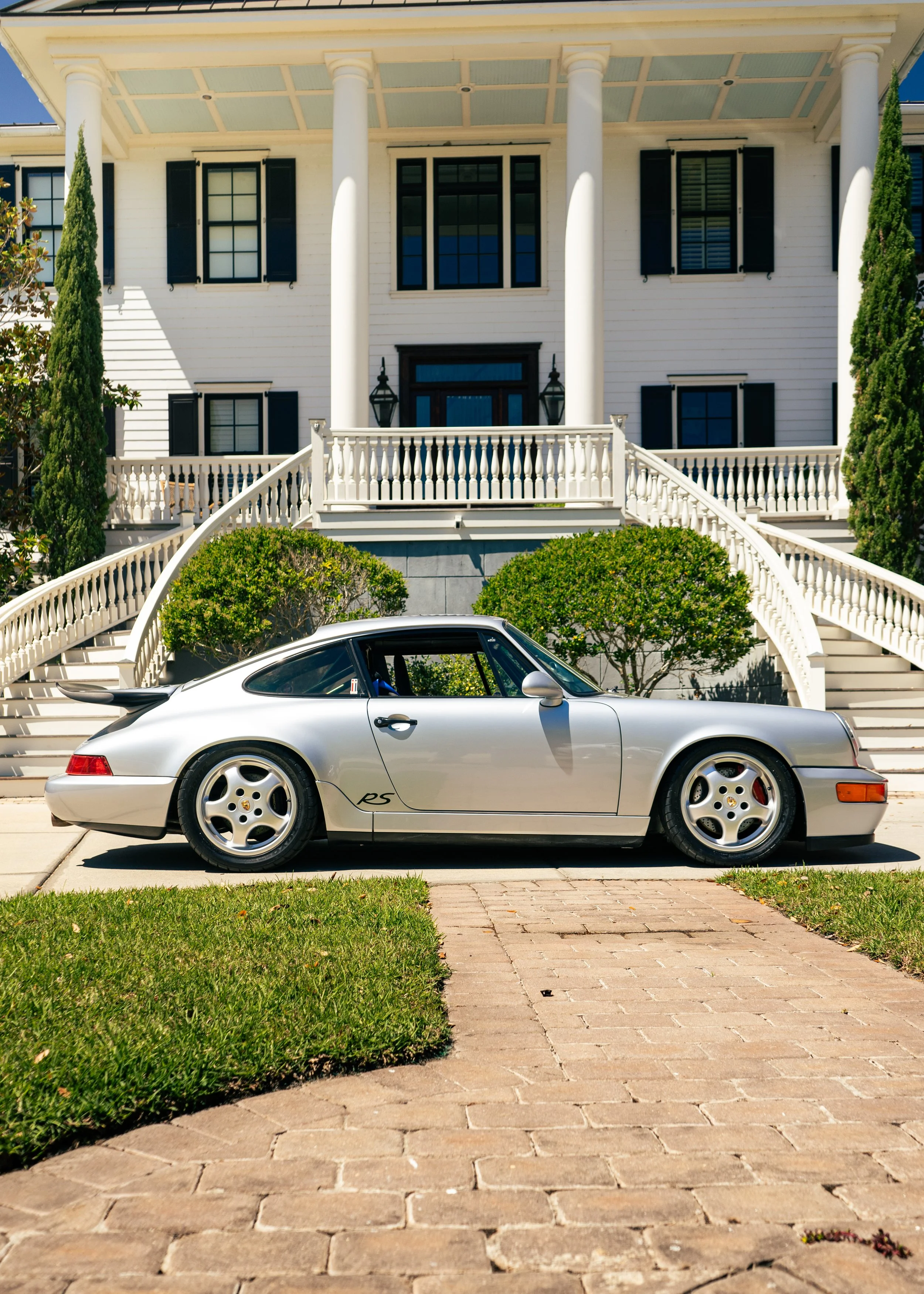 A silver Porsche sports car parked in front of a large, white house with black shutters, stairs, and greenery.