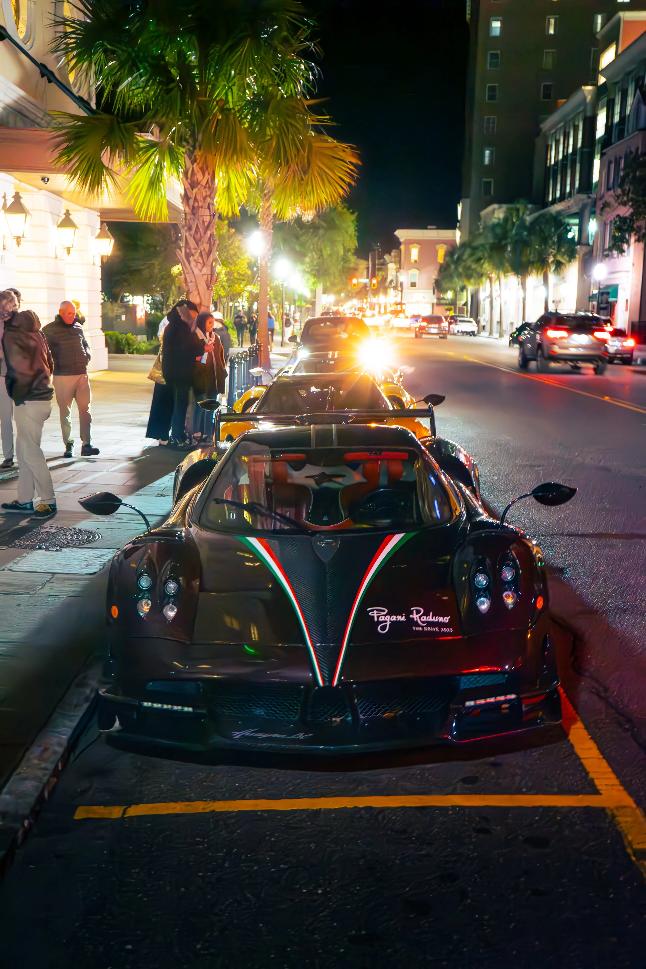 Night scene on a city street with luxury sport cars parked along the sidewalk, people gathered chatting, palm trees lining the street, buildings with lit windows, and streetlights illuminating the dark surroundings.
