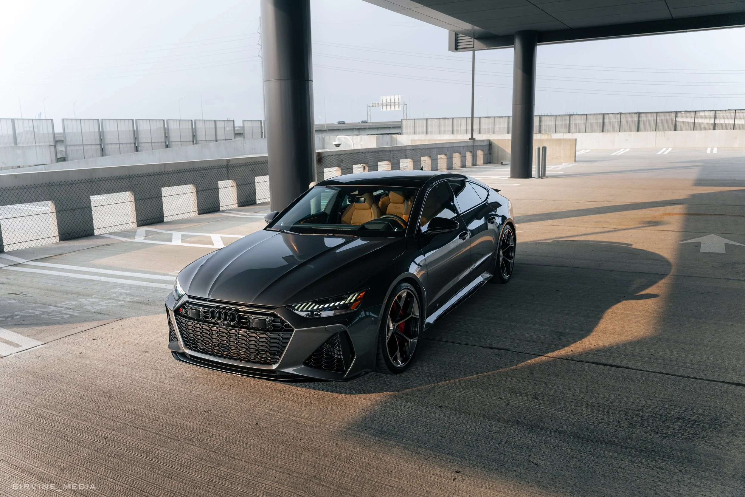 Black Audi sports car parked under an overpass on a concrete surface with a fence and highway in the background.