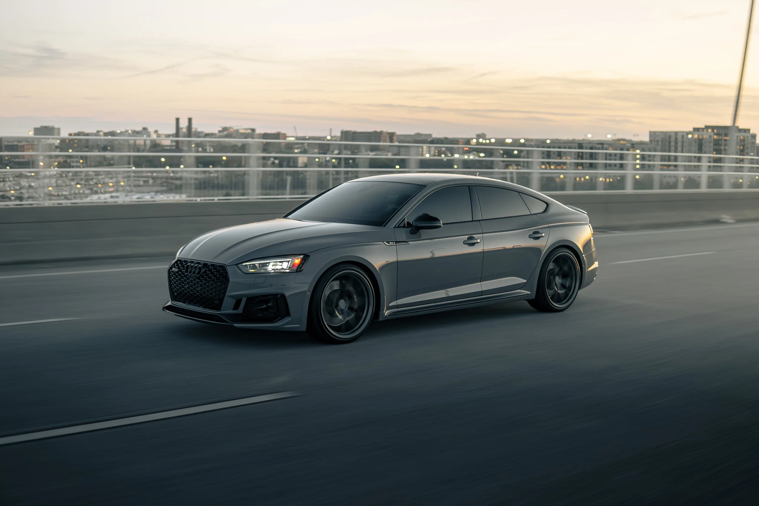 A dark gray Audi sedan driving on a highway bridge at sunset with city skyline in the background.